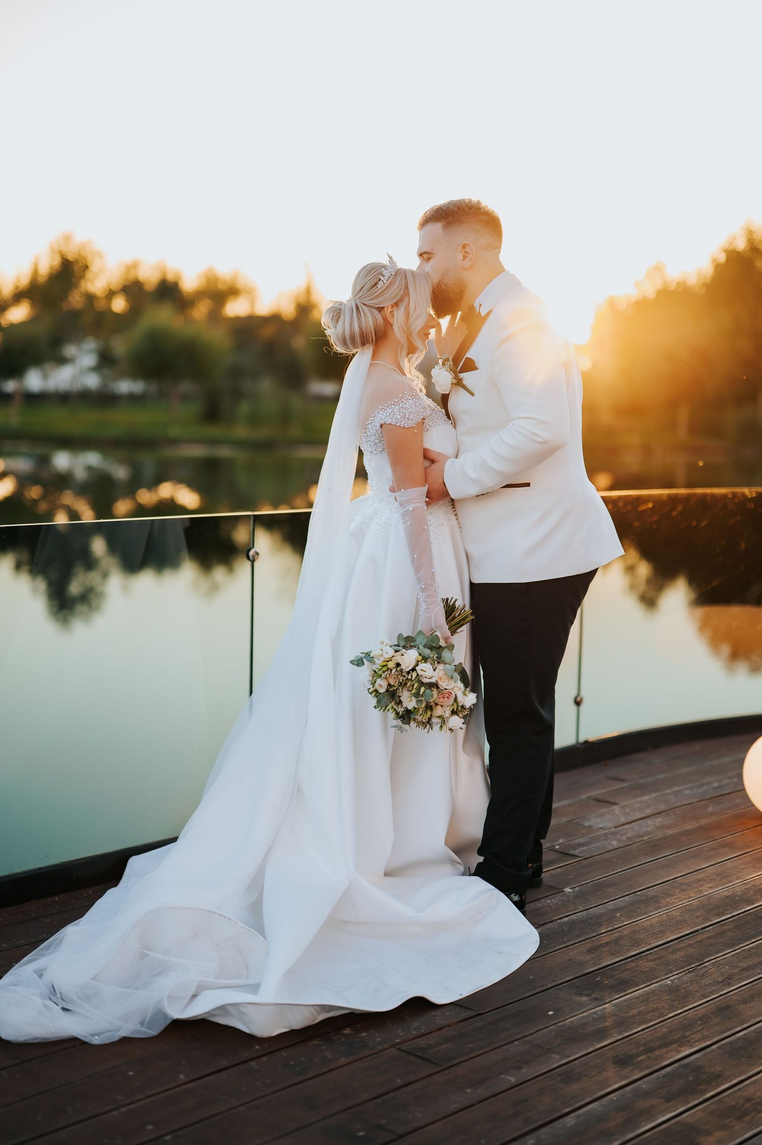 Bride and groom share a romantic sunset kiss on a dock, with golden light reflecting on a serene lake backdrop.