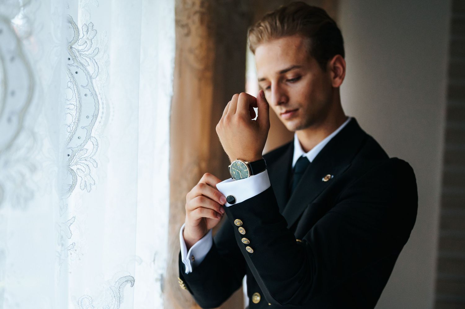 Young man in navy military uniform adjusting cufflinks by a lace curtain window in elegant setting.