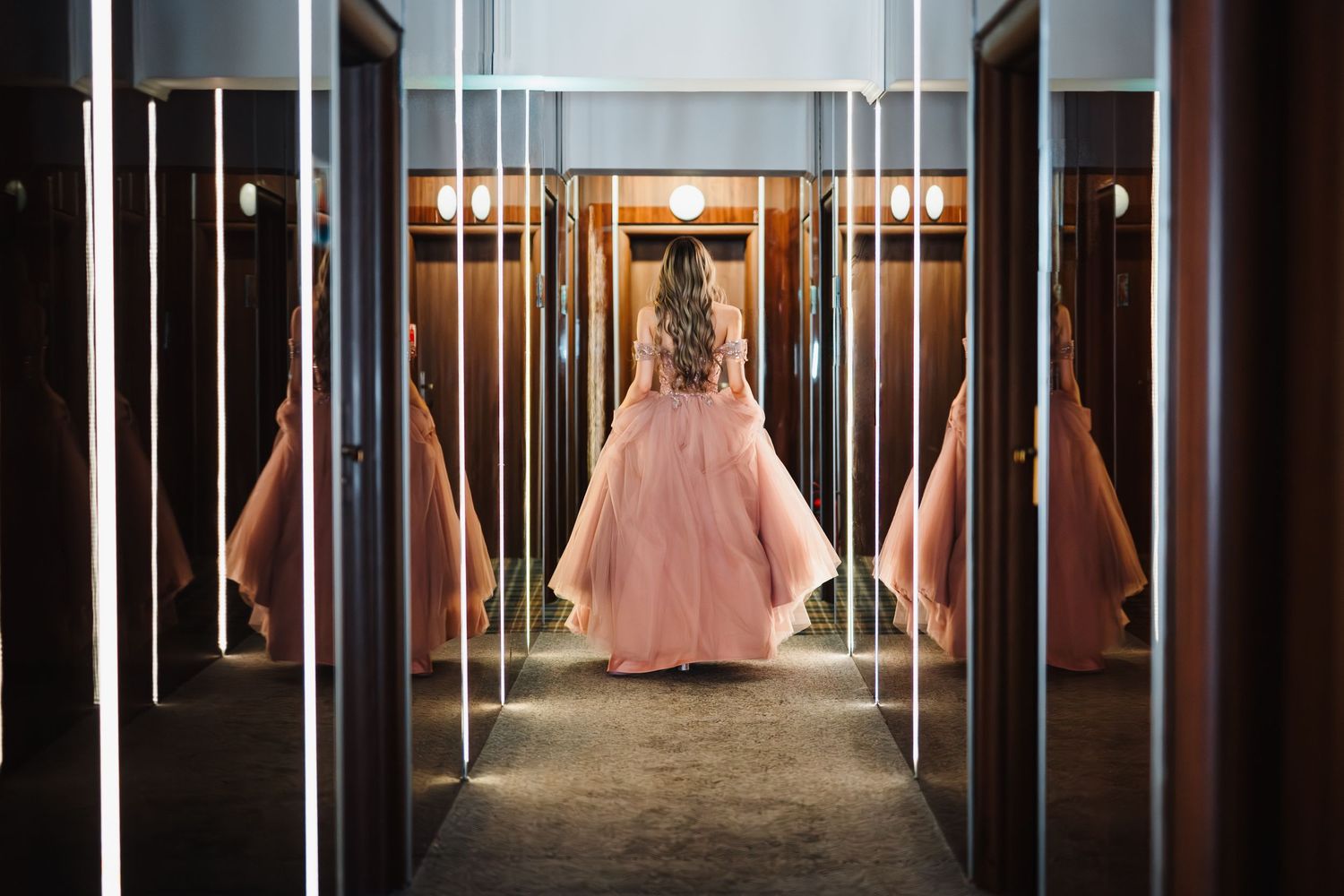 Girl in pink ball gown stands in mirrored dressing room, creating infinite reflections of her elegant dress.