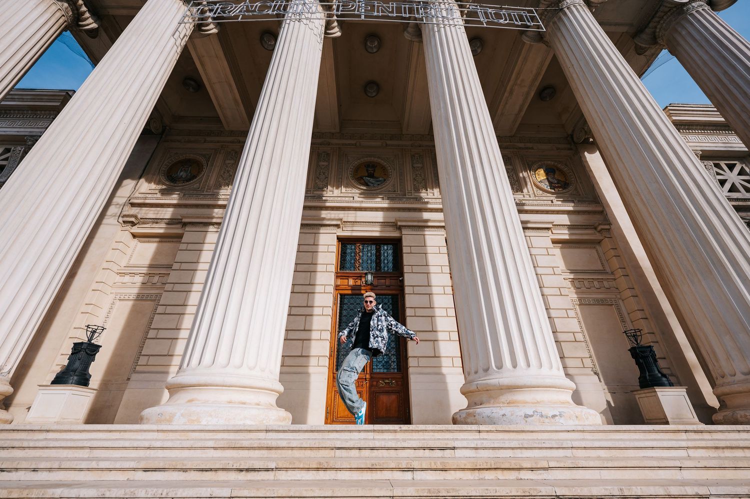 Person posing at the grand entrance of a neoclassical building with tall white columns and ornate wooden doors.
