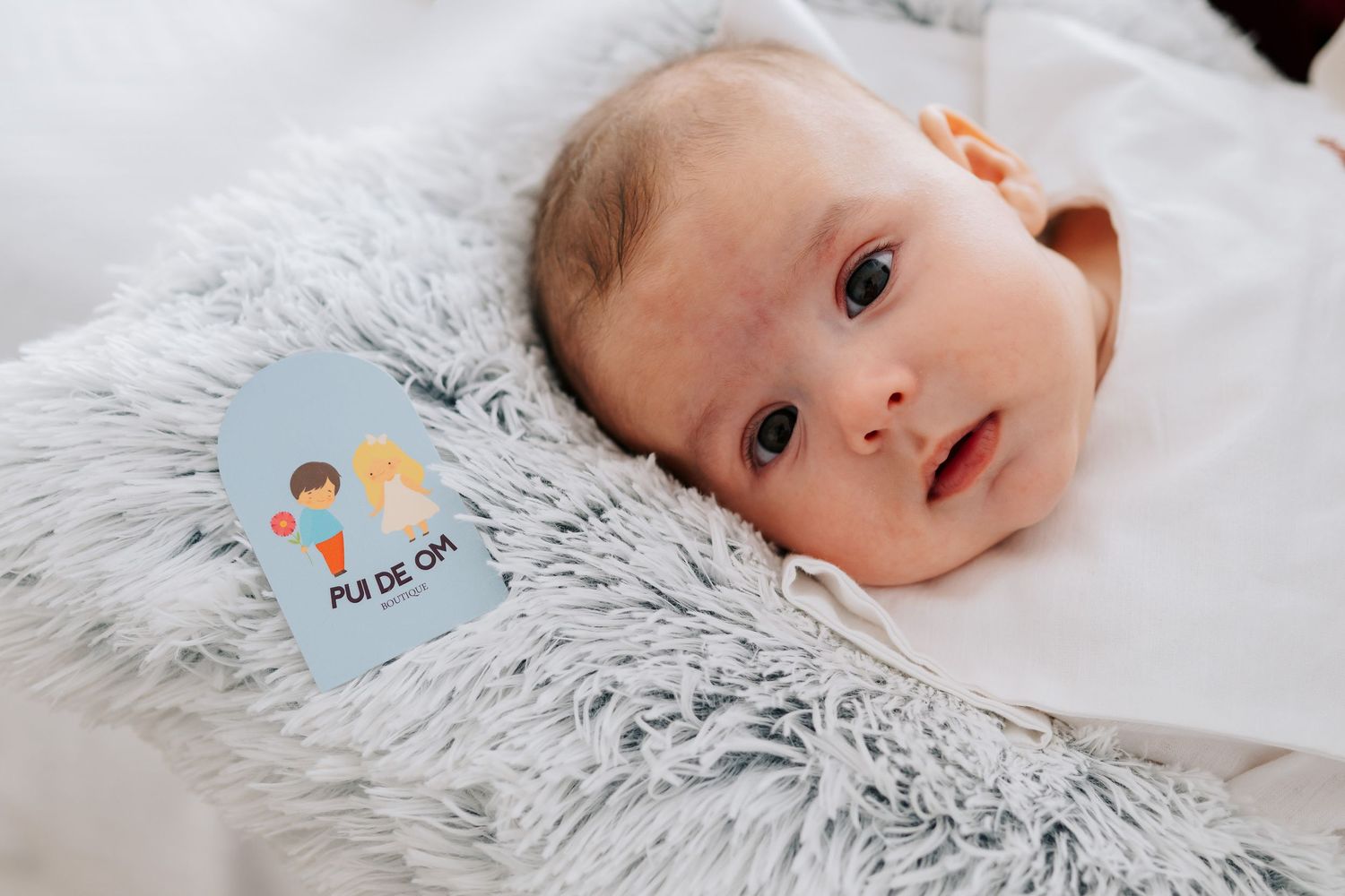 Newborn baby lying on fluffy white blanket beside a heart-shaped milestone card reading Pie de Om.