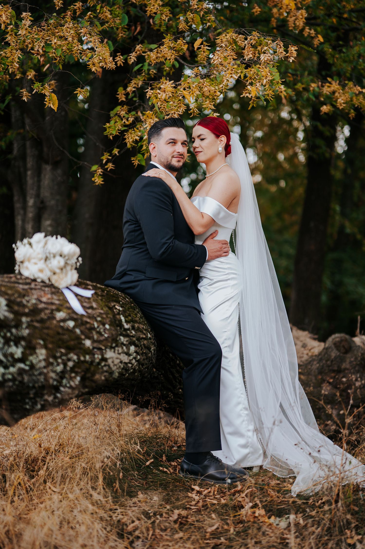 Bride in white gown and veil with groom in black suit posing romantically among rocks and autumn trees.