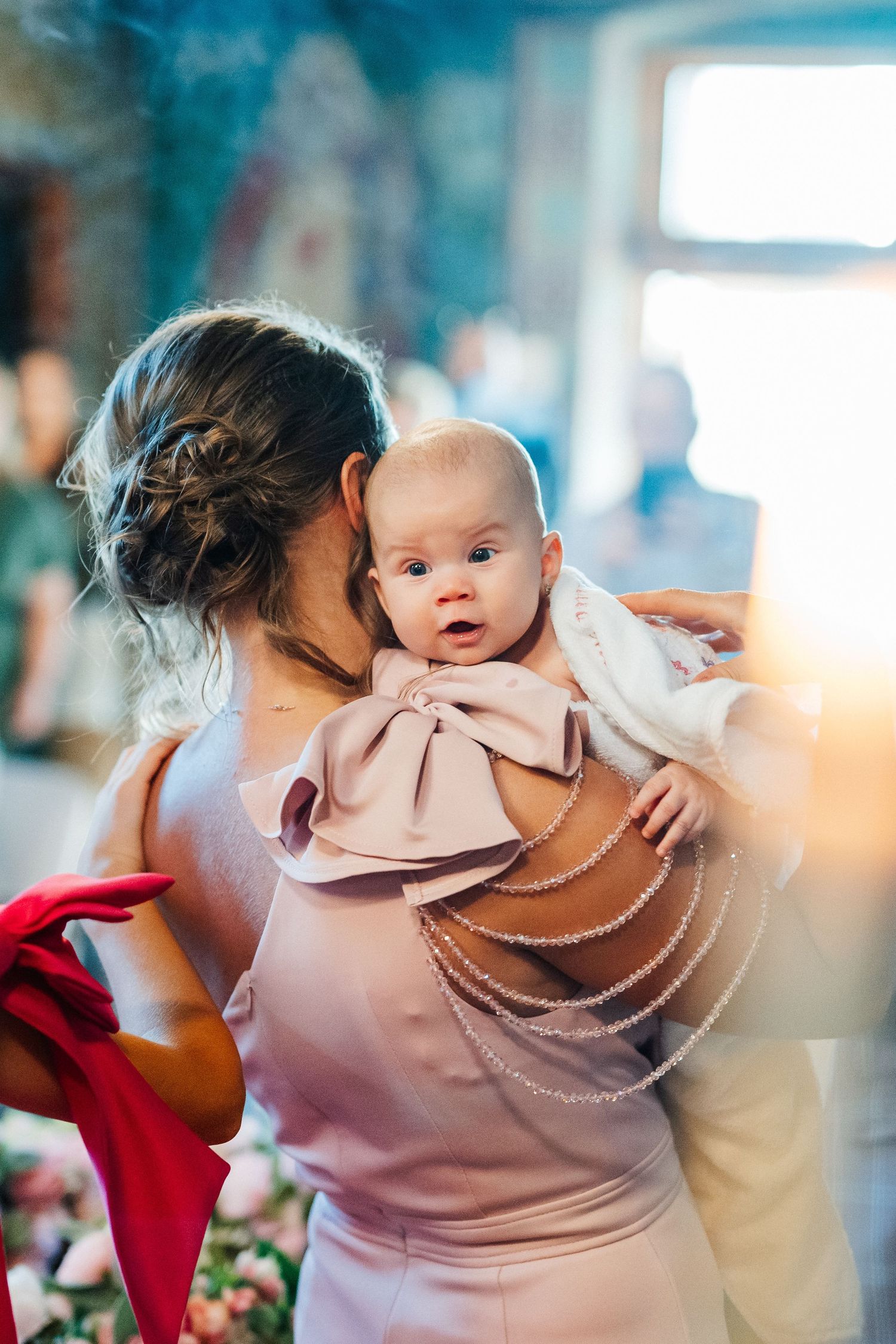 A woman in a pink dress holds a wide-eyed baby with a large bow outfit at a formal event.