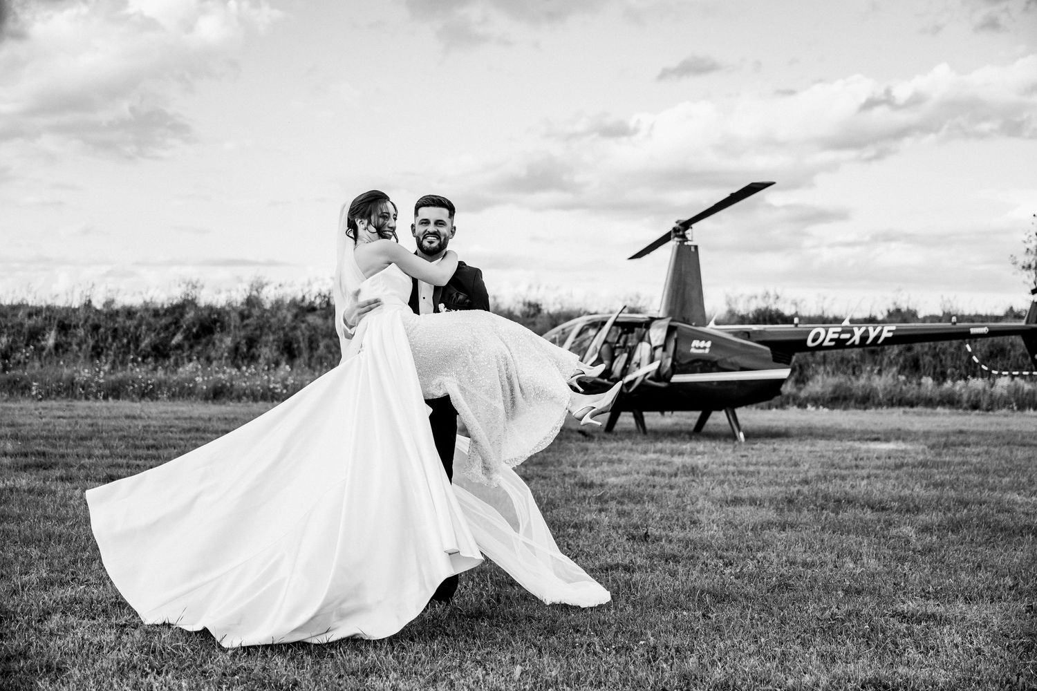 Groom carries bride in wedding dress near helicopter on grass field, black and white wedding photo.