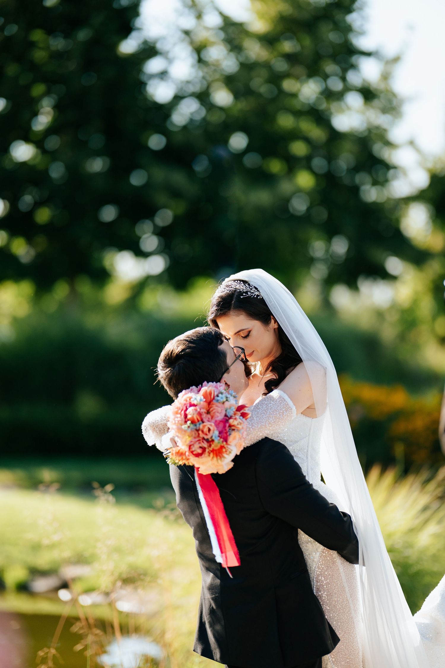 Bride and groom embracing outdoors, bride holding colorful bouquet with pink ribbon, wearing white lace gown and veil.