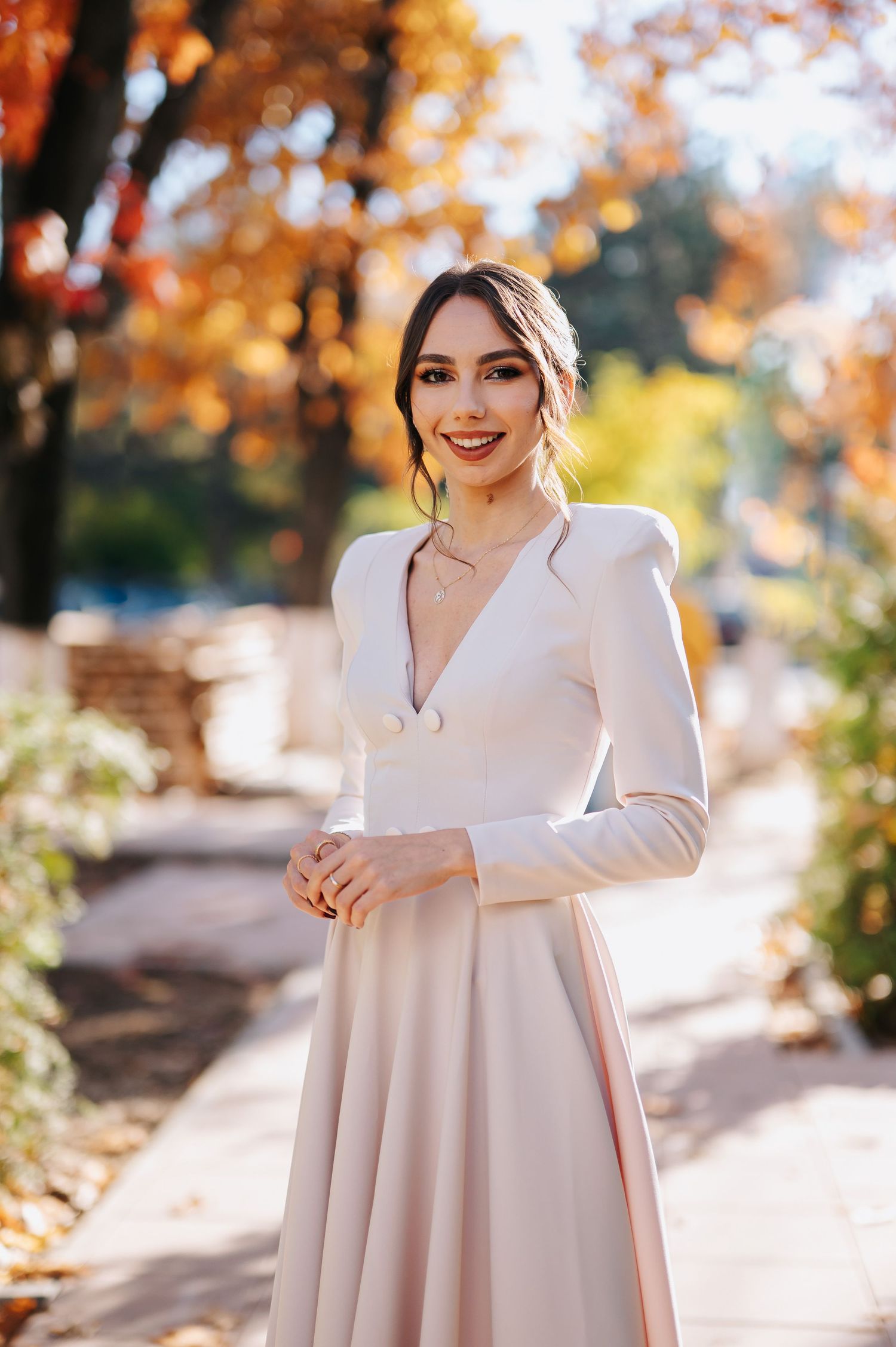 Elegant woman in blush pink long-sleeve dress smiling outdoors among autumn foliage on a sunny day.