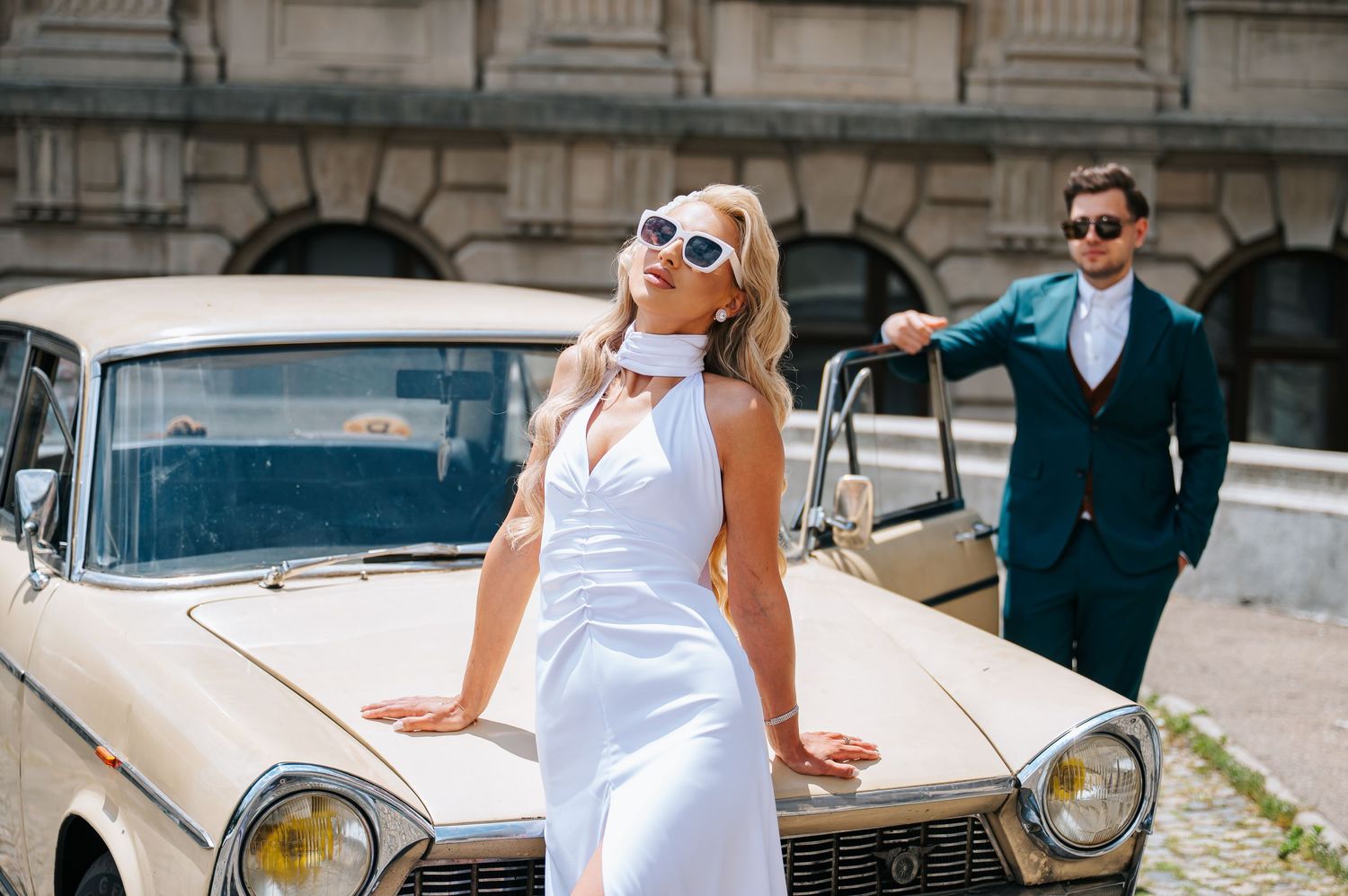 Stylish bride in white dress poses by vintage car with groom in teal suit.