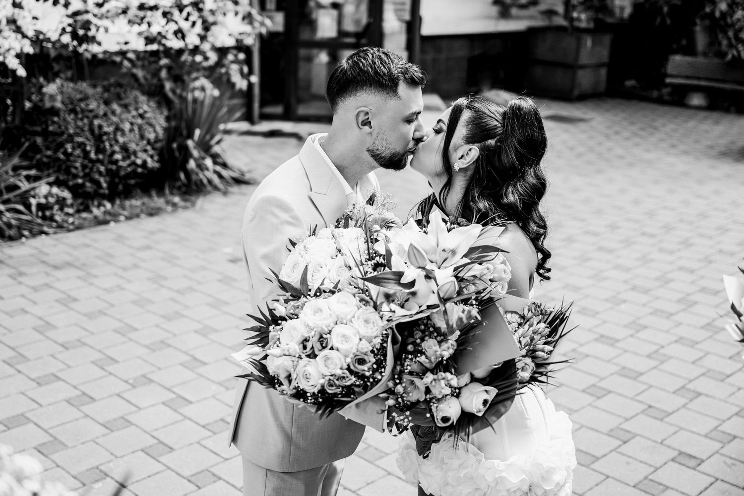 Bride and groom share romantic kiss outdoors holding large floral bouquet in black and white wedding photo.