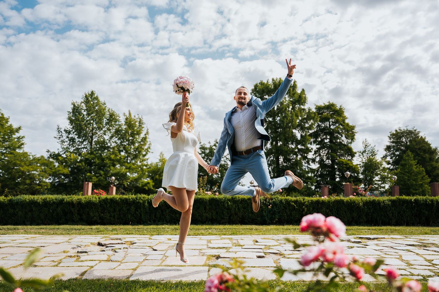 Joyful newlyweds jumping together in sunny garden with blue sky and green hedges behind them.