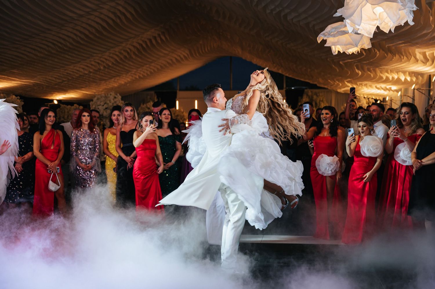 Groom lifting bride on foggy dance floor at wedding reception with guests in red and gold gowns.