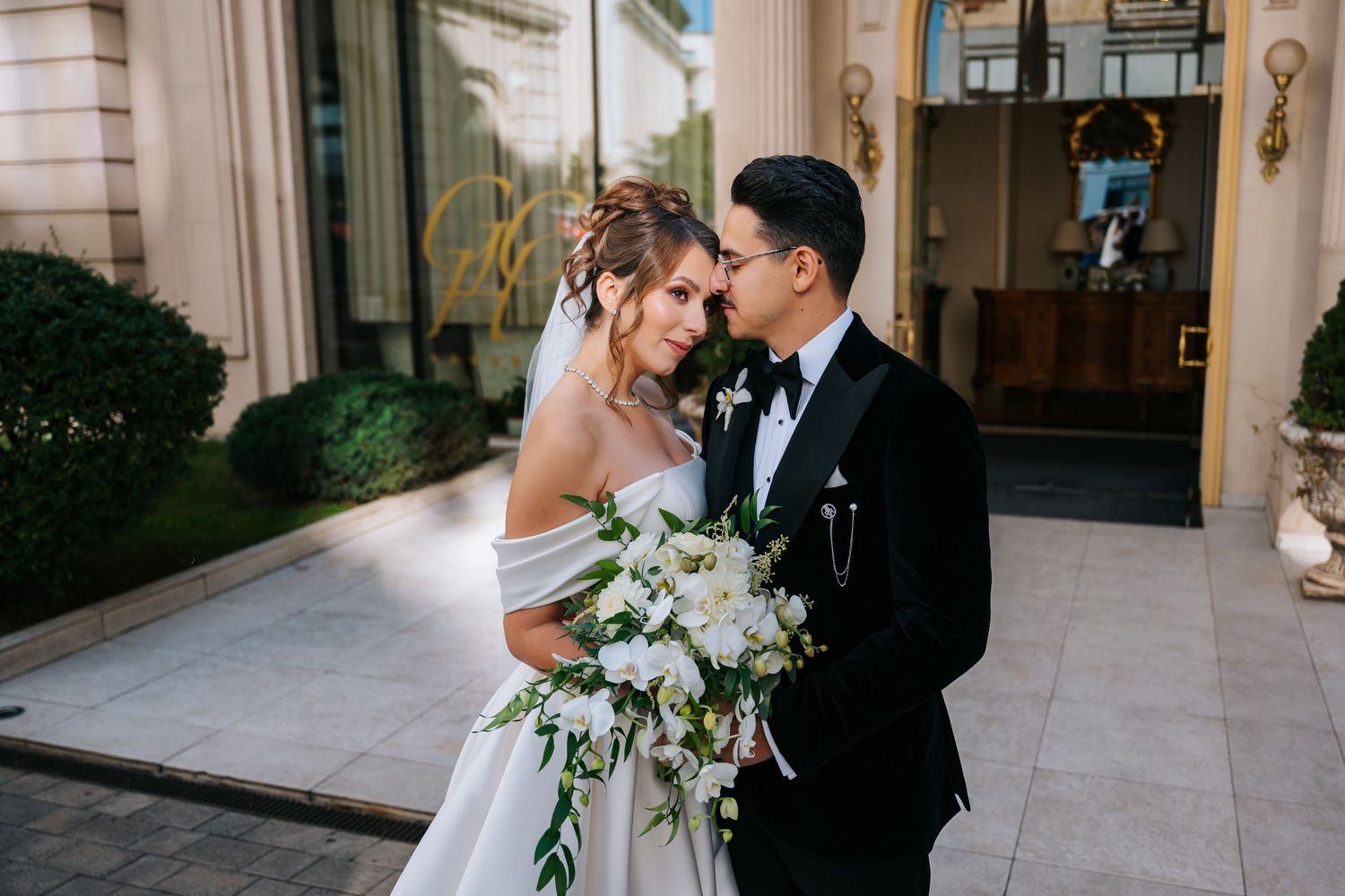 Elegant bride and groom share romantic moment outside luxury venue, bride holding white floral bouquet in stunning wedding gown.