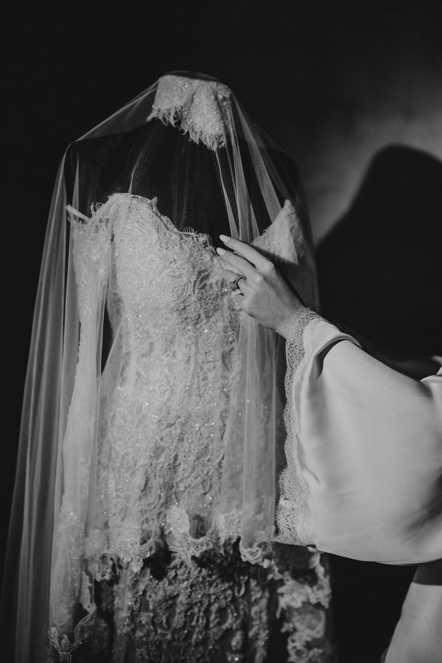 Black and white close-up of bride touching lace wedding dress with veil on mannequin.
