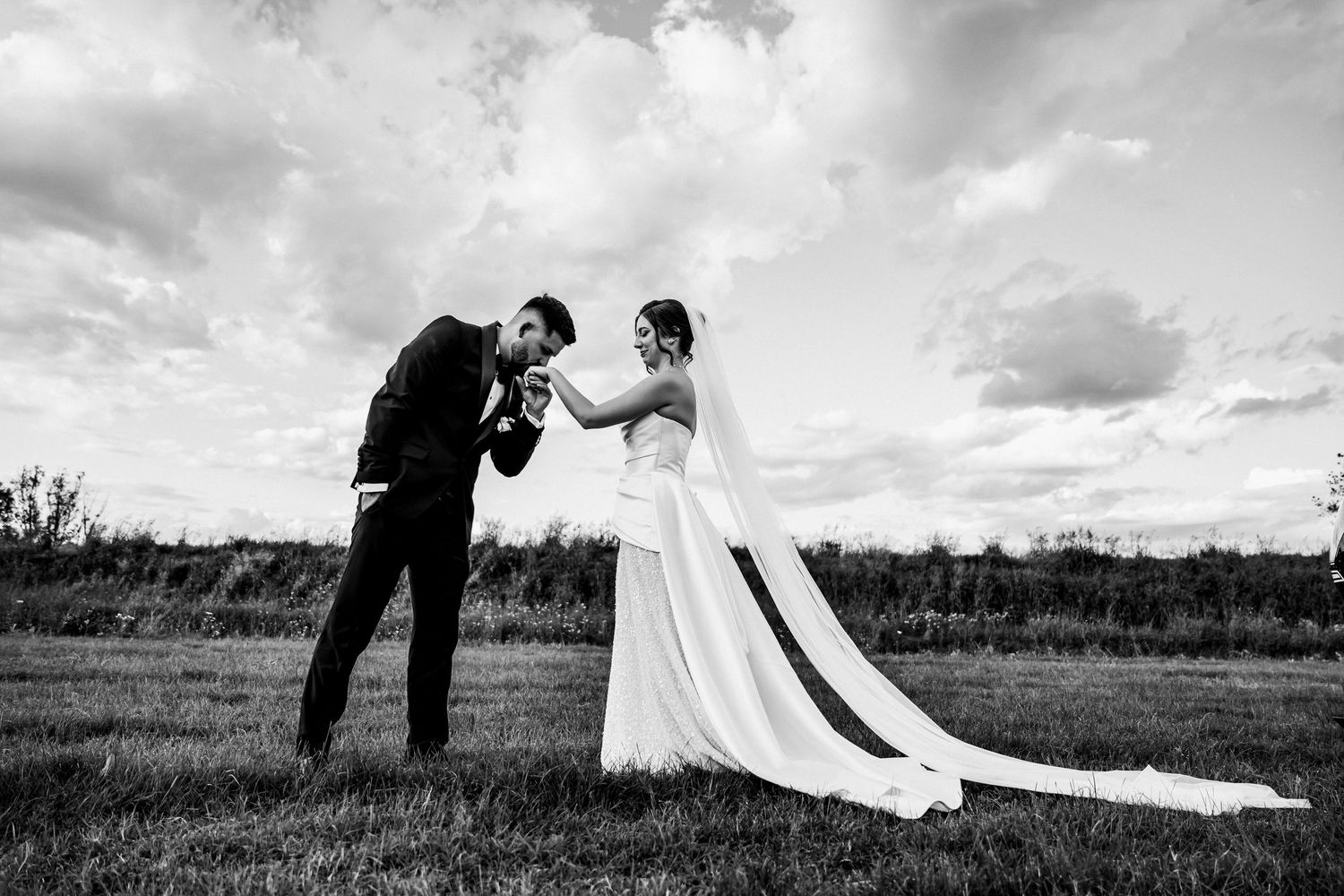 Black and white photo of a bride and groom sharing a romantic moment in a field under a cloudy sky.