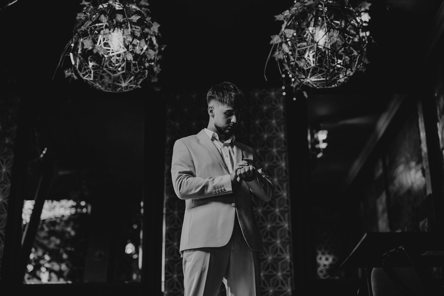 Black and white photo of a groom in a light suit adjusting his cufflinks under ornate chandeliers.