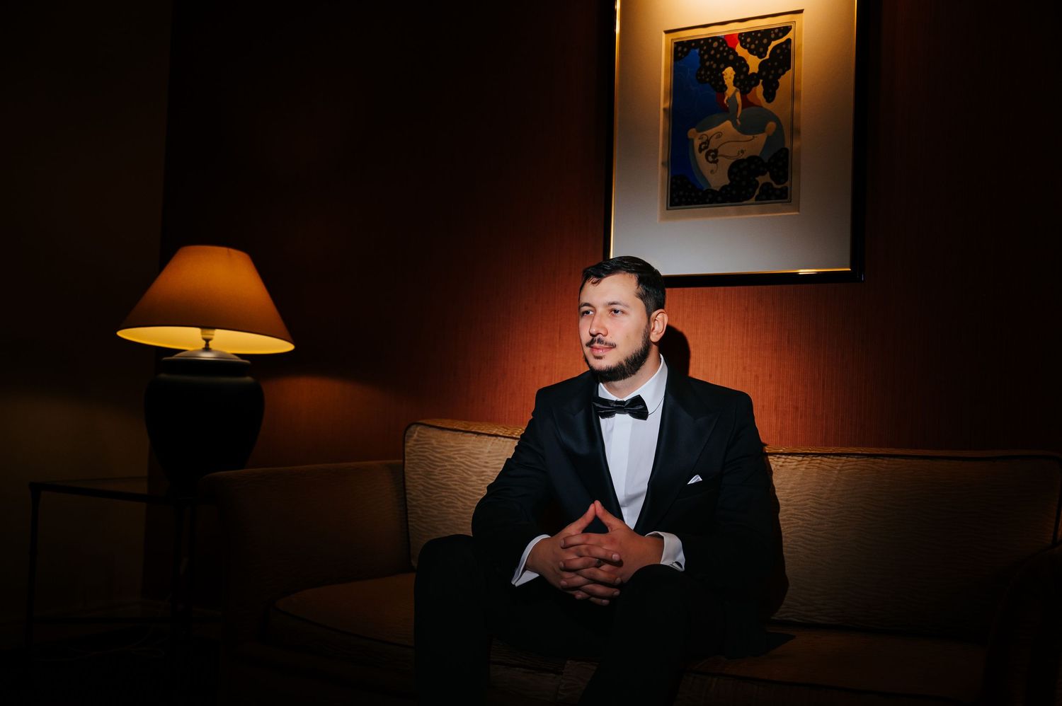 Elegant groom in black tuxedo and bow tie sitting on a sofa in a dimly lit room with a lamp and artwork.
