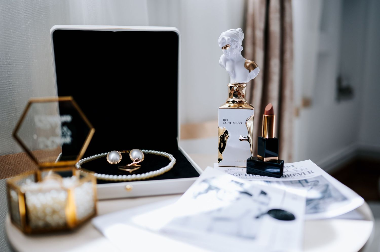 Elegant bridal accessories including pearl jewelry, gold lipstick, angel perfume bottle, and a velvet jewelry box on a white table.