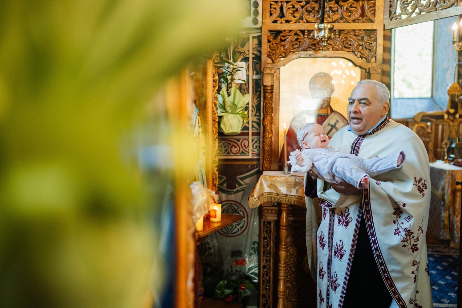Orthodox priest holding infant during baptism ceremony inside ornate church with golden iconostasis.