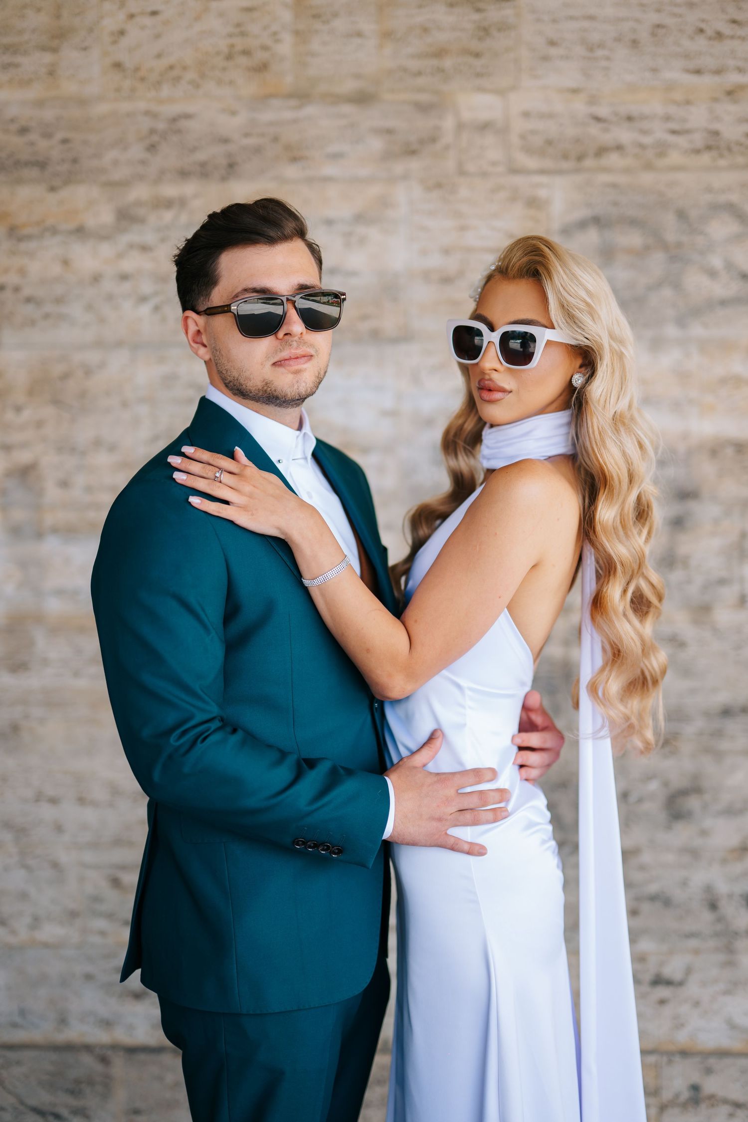 Stylish couple in sunglasses posing together, man in teal suit and woman in elegant white dress against stone wall.