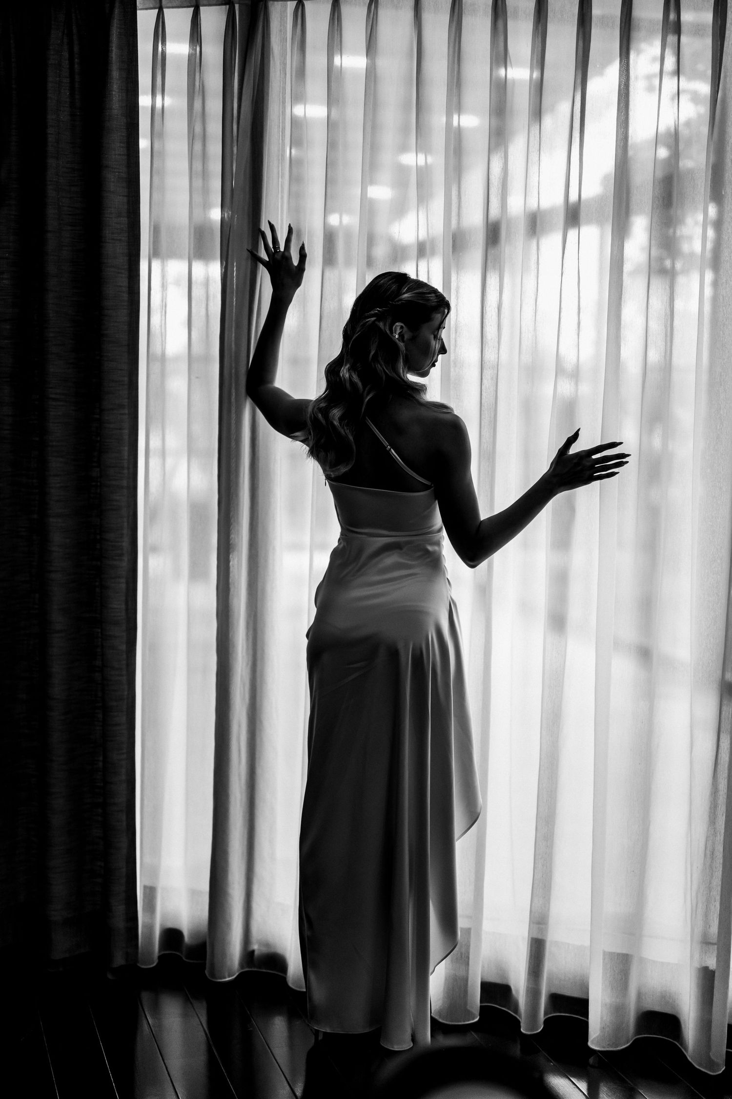 Elegant bride silhouetted in black and white standing before sheer curtained window.