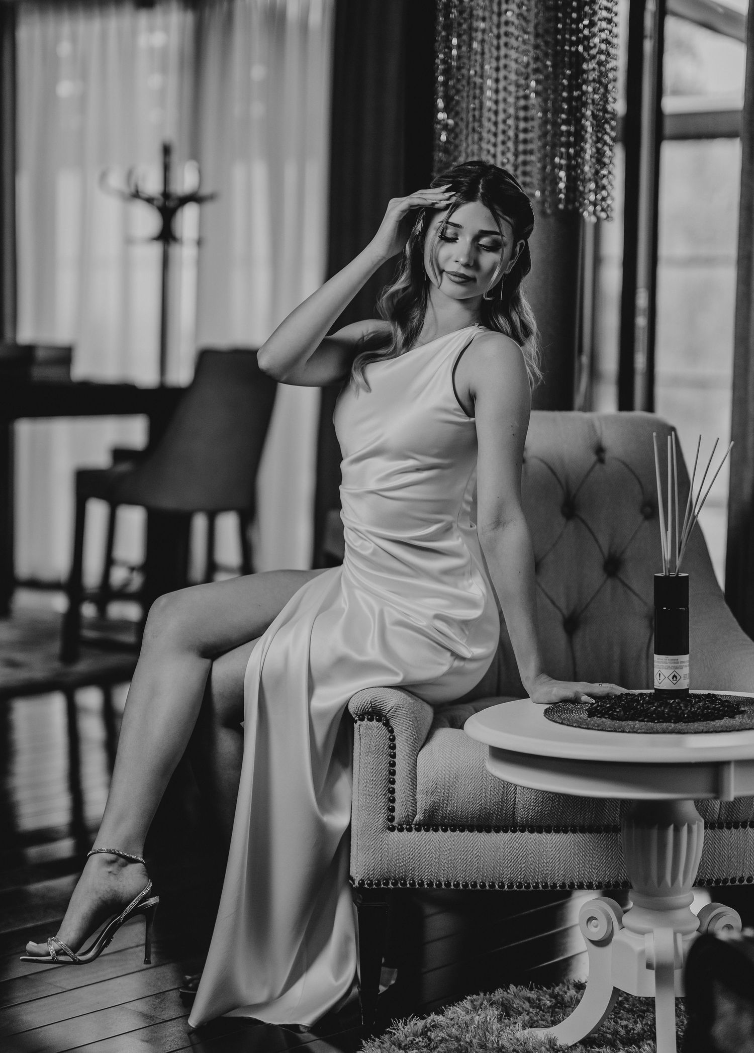 Elegant woman in white satin slip dress seated in tufted chair, black and white luxury interior photo.