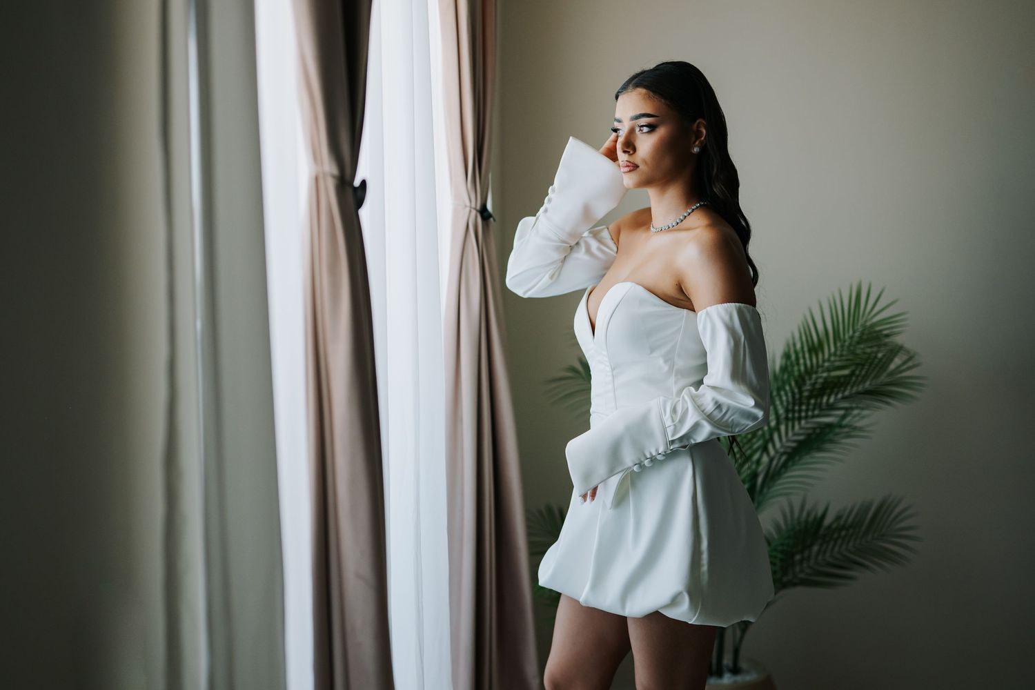 A young woman in a white off-shoulder mini dress stands by a curtained window with a palm plant nearby.