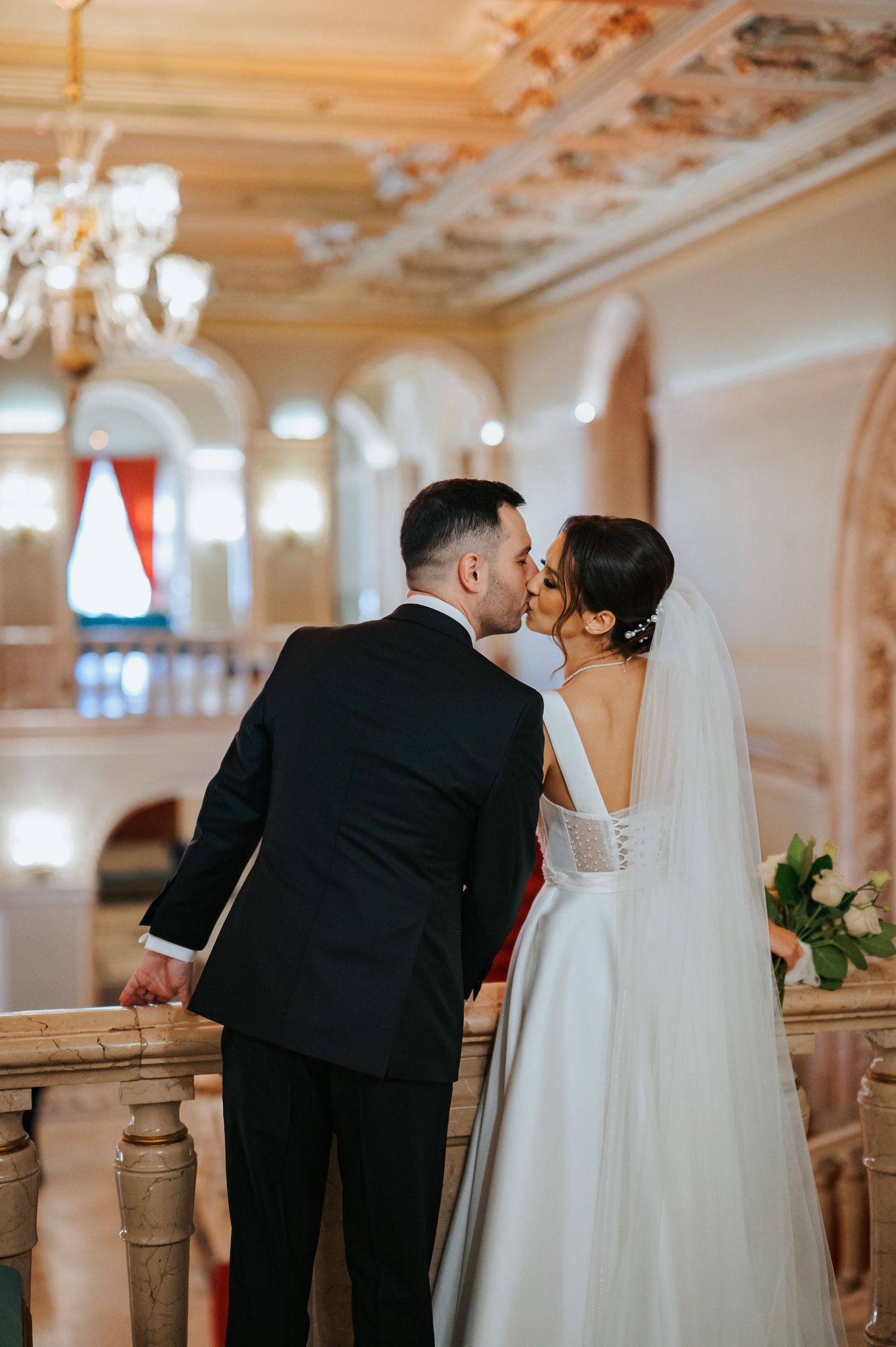Bride and groom share a kiss on an ornate balcony in an elegant ballroom, bride wearing a white gown and veil.