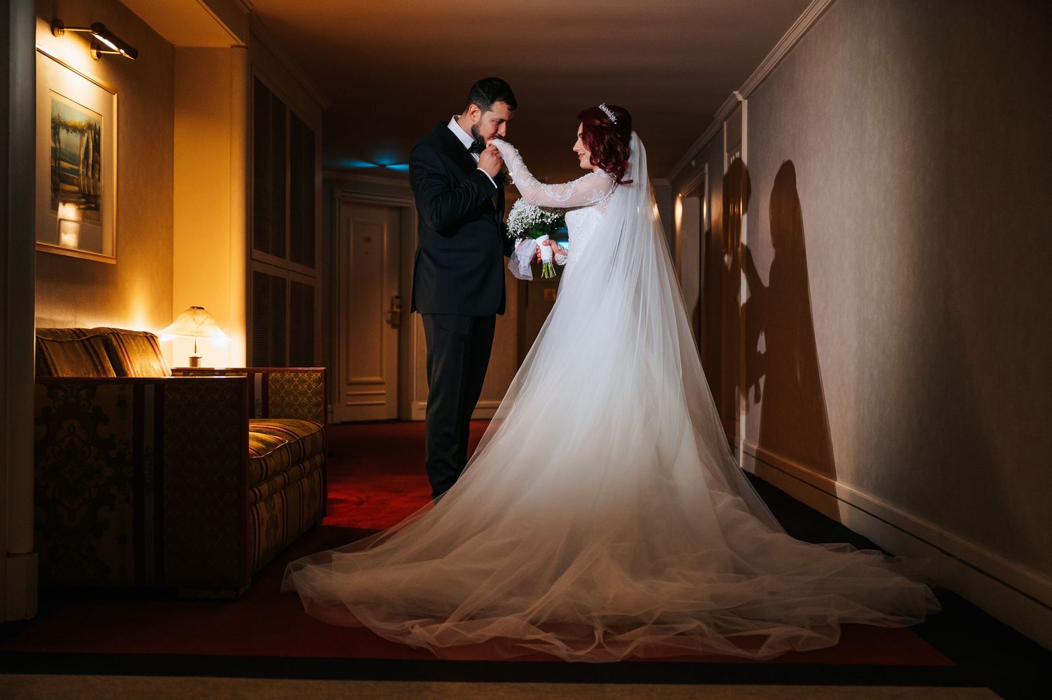 A bride and groom share a romantic moment in a warmly lit hotel hallway, her white gown casting a dramatic shadow.