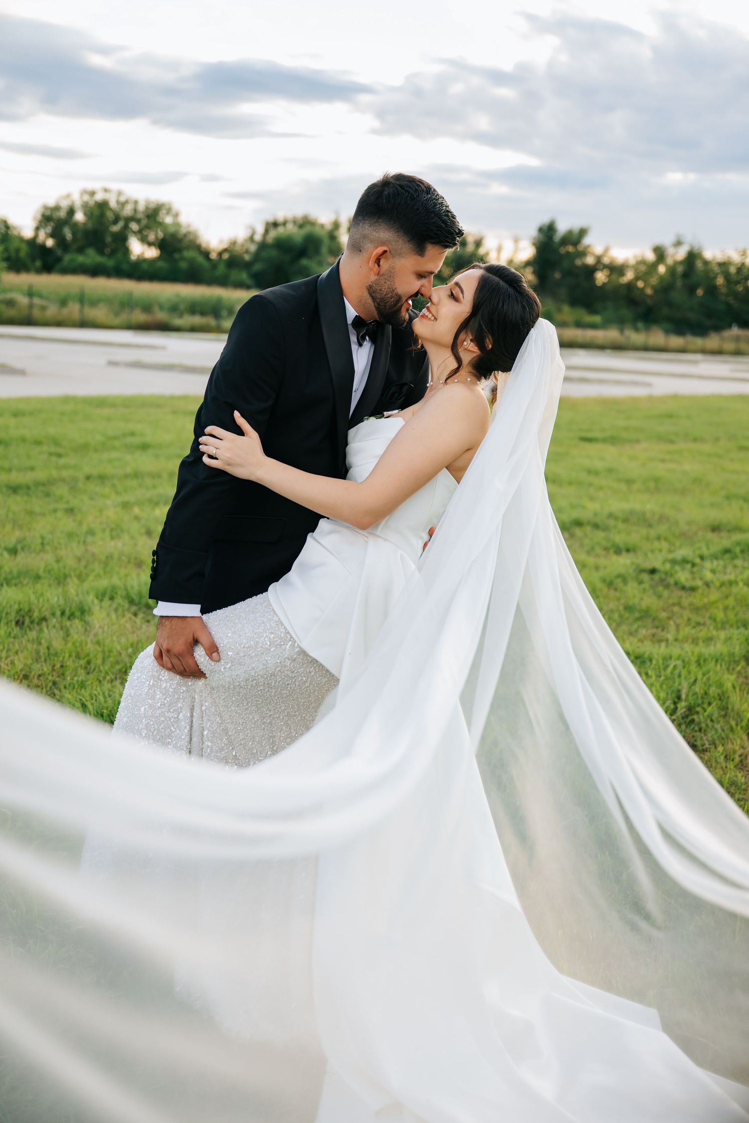 Bride and groom embrace in a green field, her veil flowing in the wind, both smiling in elegant wedding attire.