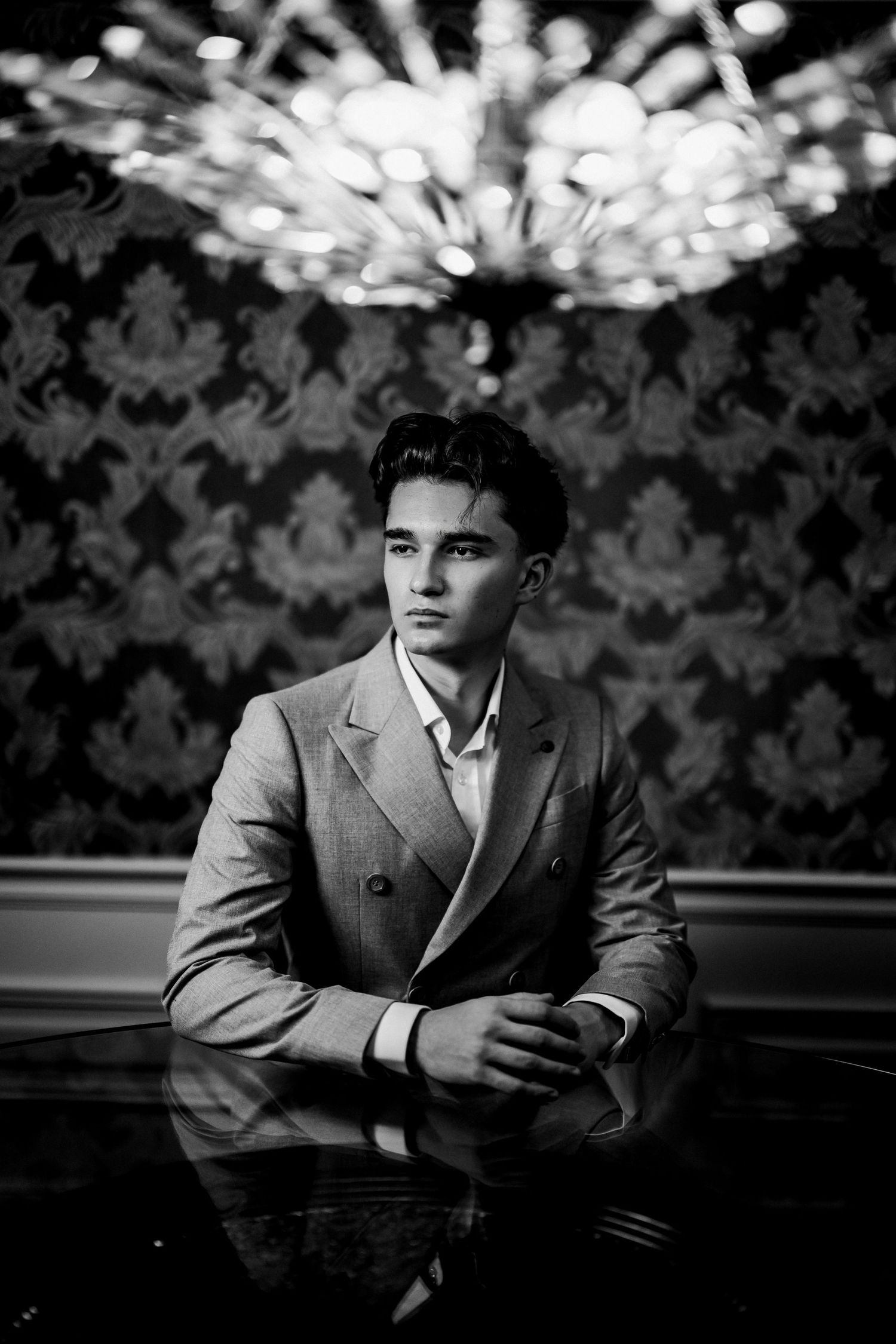 Black and white portrait of a young man in a double-breasted suit seated at a table under a chandelier.