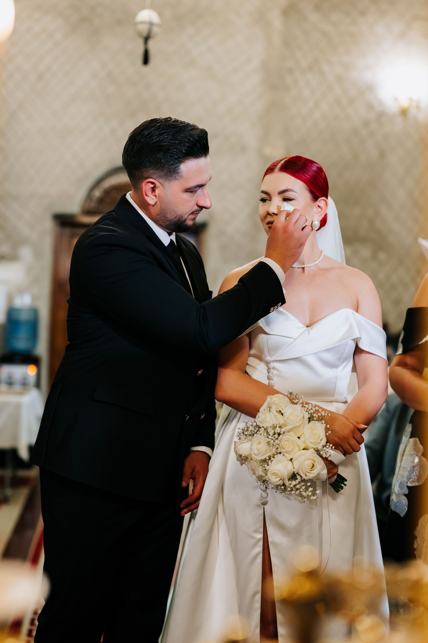 Groom in black suit gently wipes tears from bride's cheek during emotional wedding ceremony.