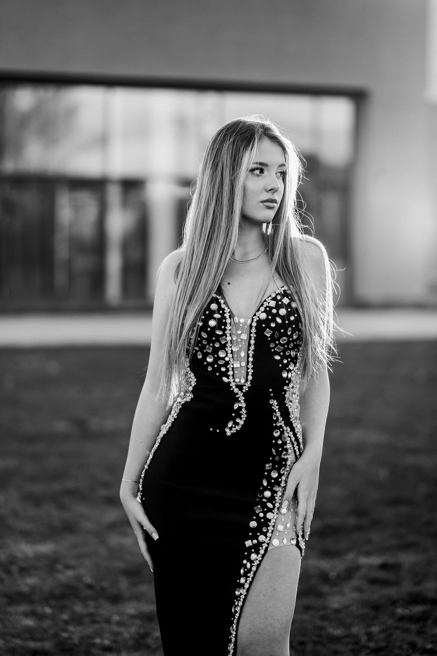 Black and white photo of a young woman in an elegant beaded gown posing outdoors near a modern building.