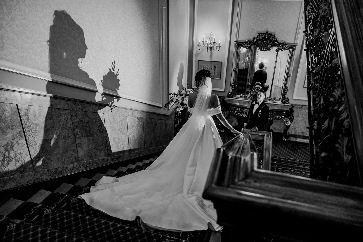 Black and white artistic photo of bride descending grand staircase with flowing satin gown.