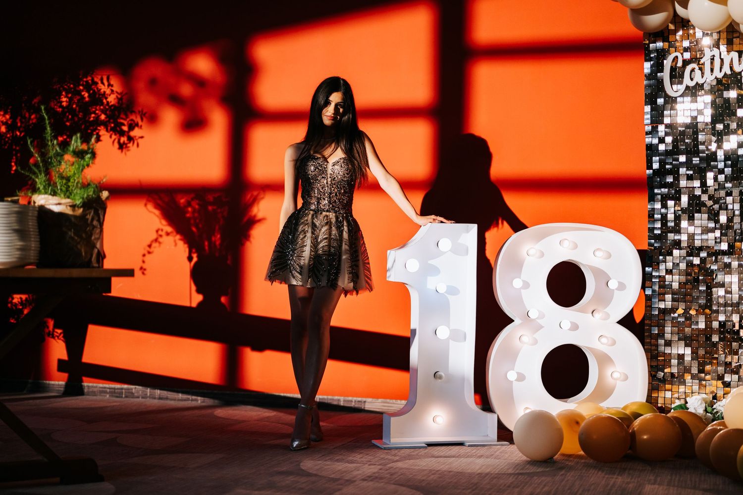Young woman in black dress posing next to illuminated number 18 sign at a birthday party celebration.