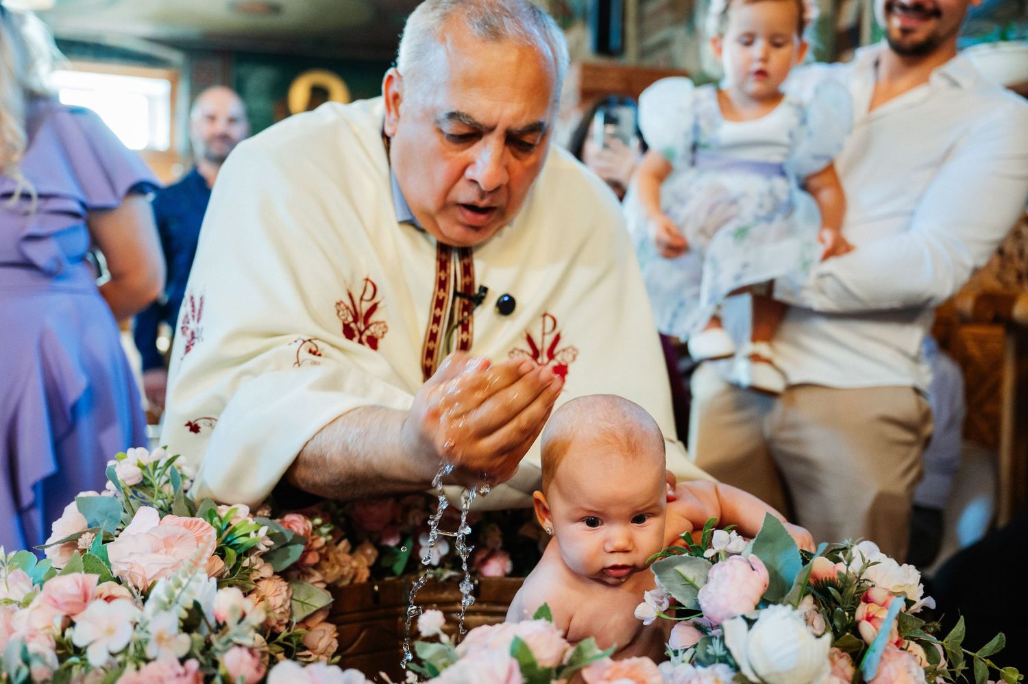Priest performing a baby baptism ceremony with floral decorations and a rosary in a church setting.