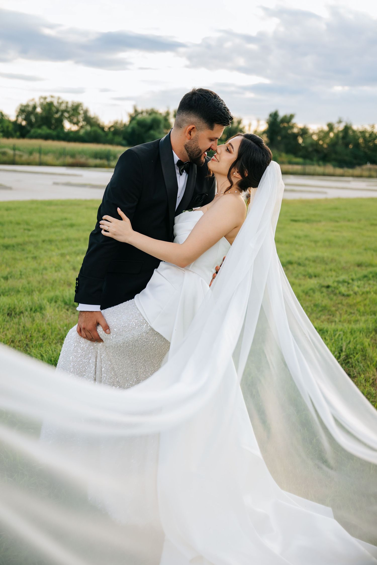 Bride and groom embracing outdoors, her flowing veil billowing in the wind against a lush green field backdrop.