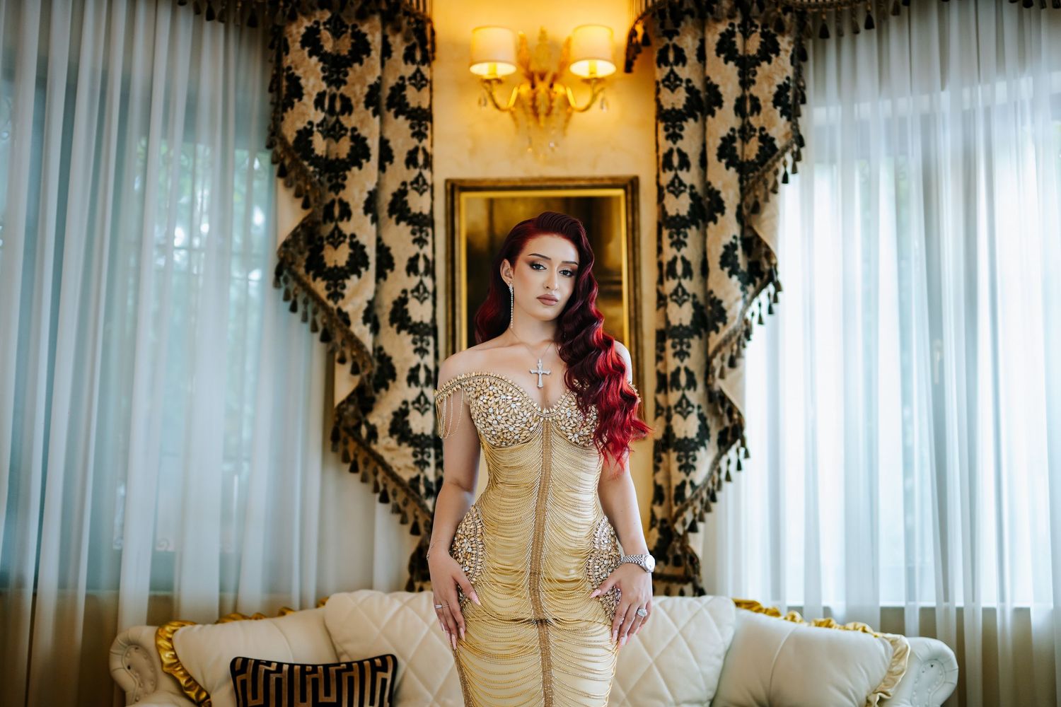 Woman in gold sequin gown with red hair posing elegantly on a cream sofa in a luxurious decorated room.