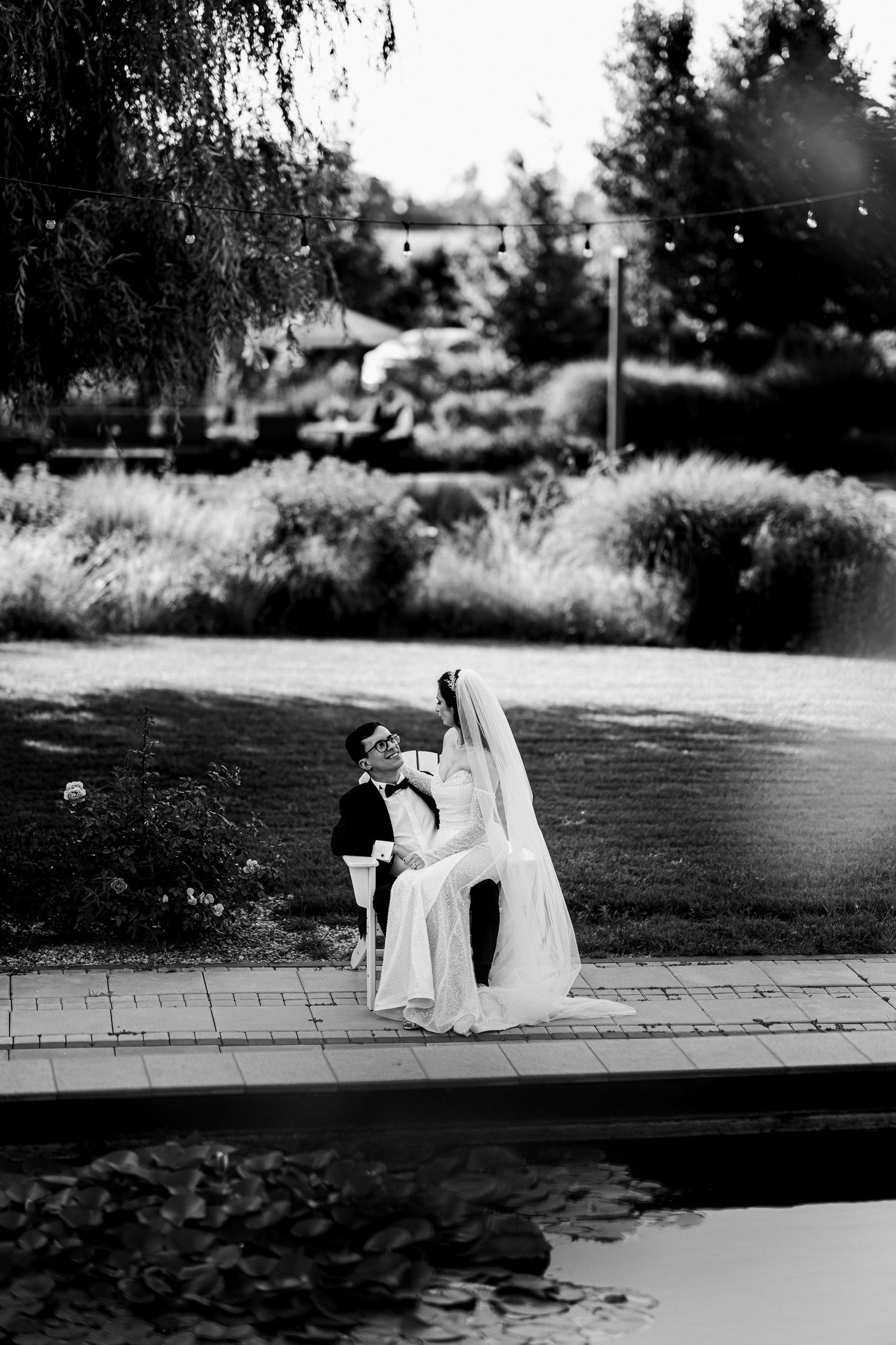 Black and white photo of bride and groom embracing by a reflective pond surrounded by lush gardens on their wedding day.