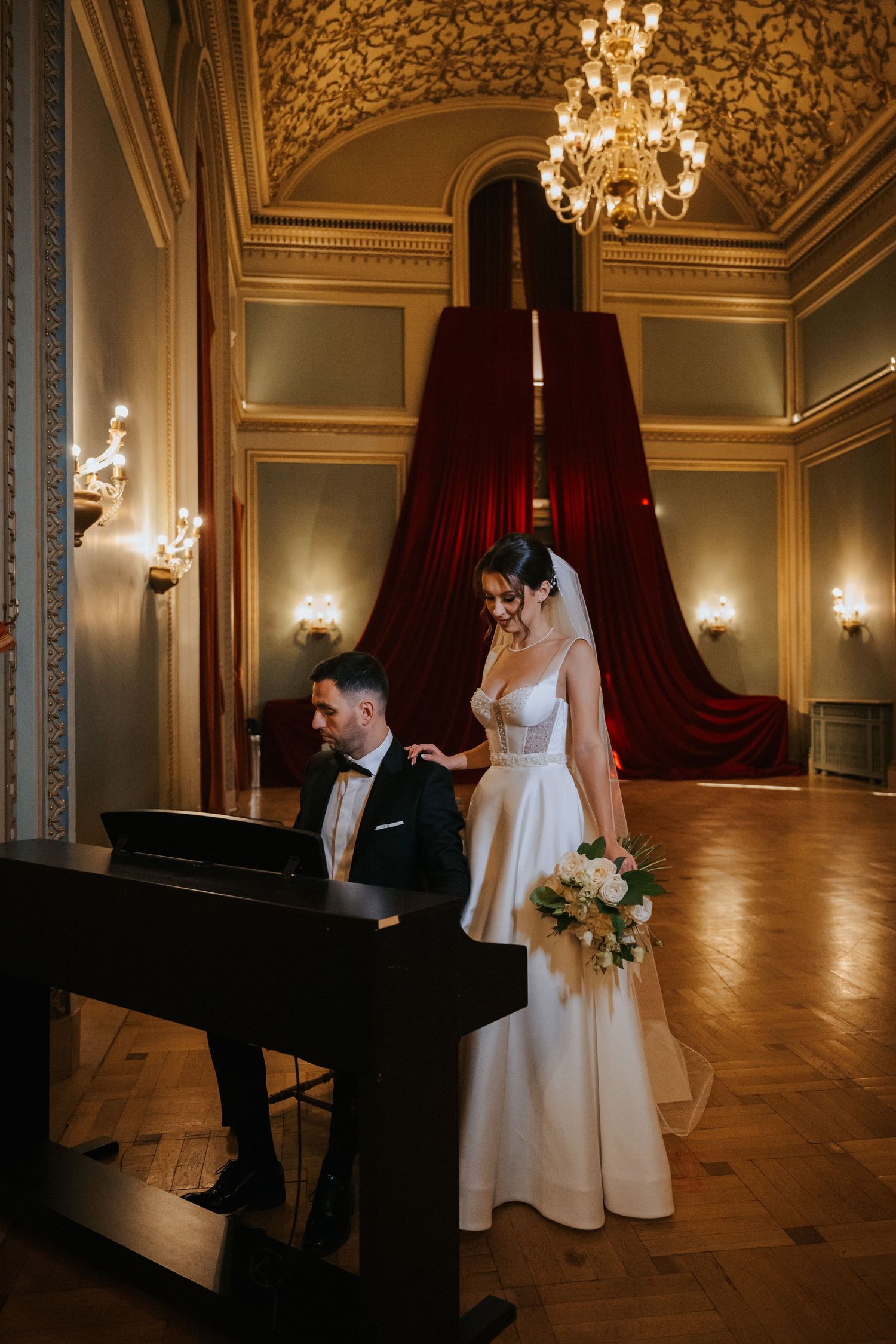 Groom playing piano in ornate ballroom while bride in white gown holds bouquet, elegant chandelier overhead.