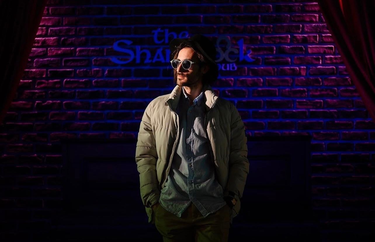 Man in jacket posing dramatically under colorful stage lights against brick wall backdrop.