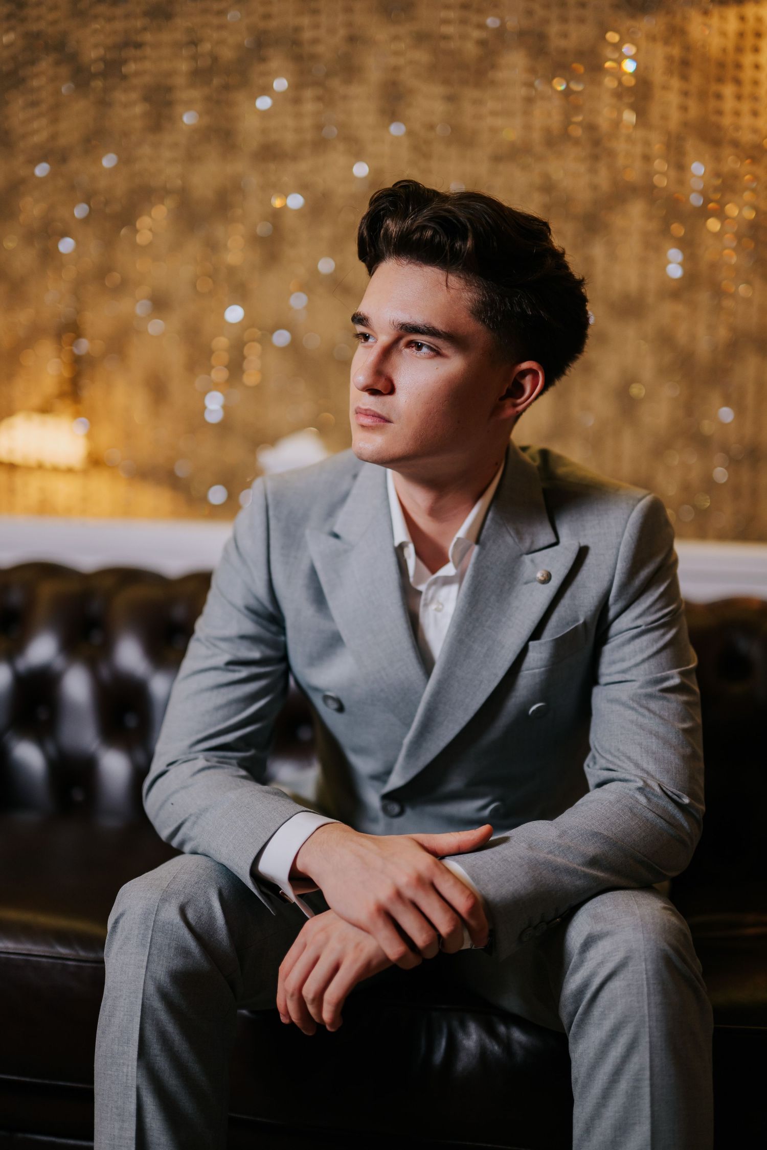 Young man in gray suit sitting on leather sofa with golden bokeh background, looking thoughtfully to the side.