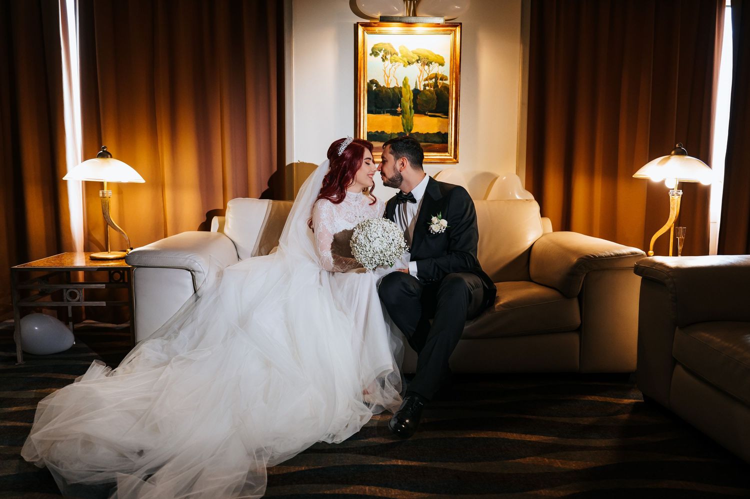 Bride and groom share a romantic kiss while seated on a couch in an elegantly lit hotel suite.
