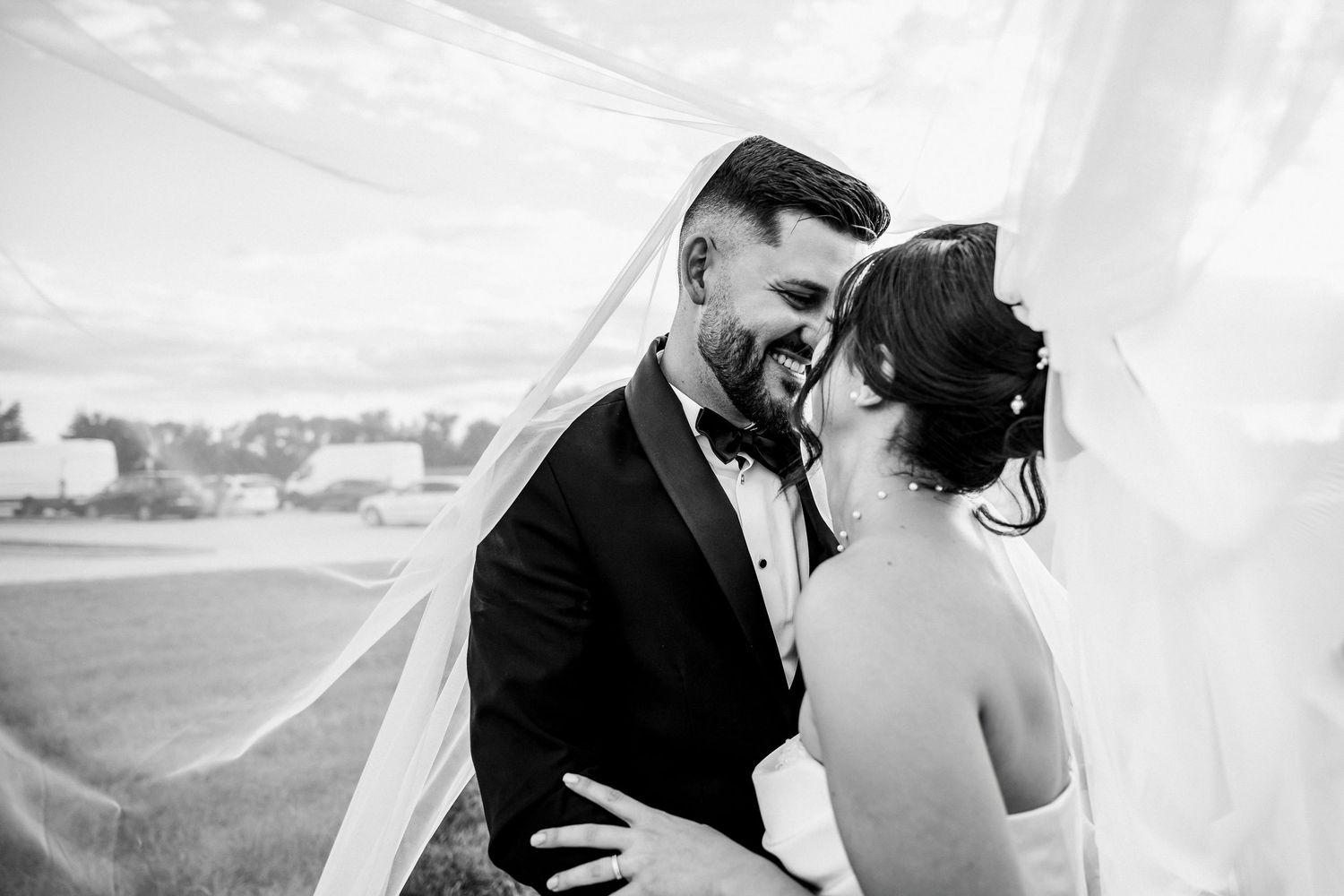 Black and white wedding photo of bride and groom sharing romantic moment under flowing veil outdoors.