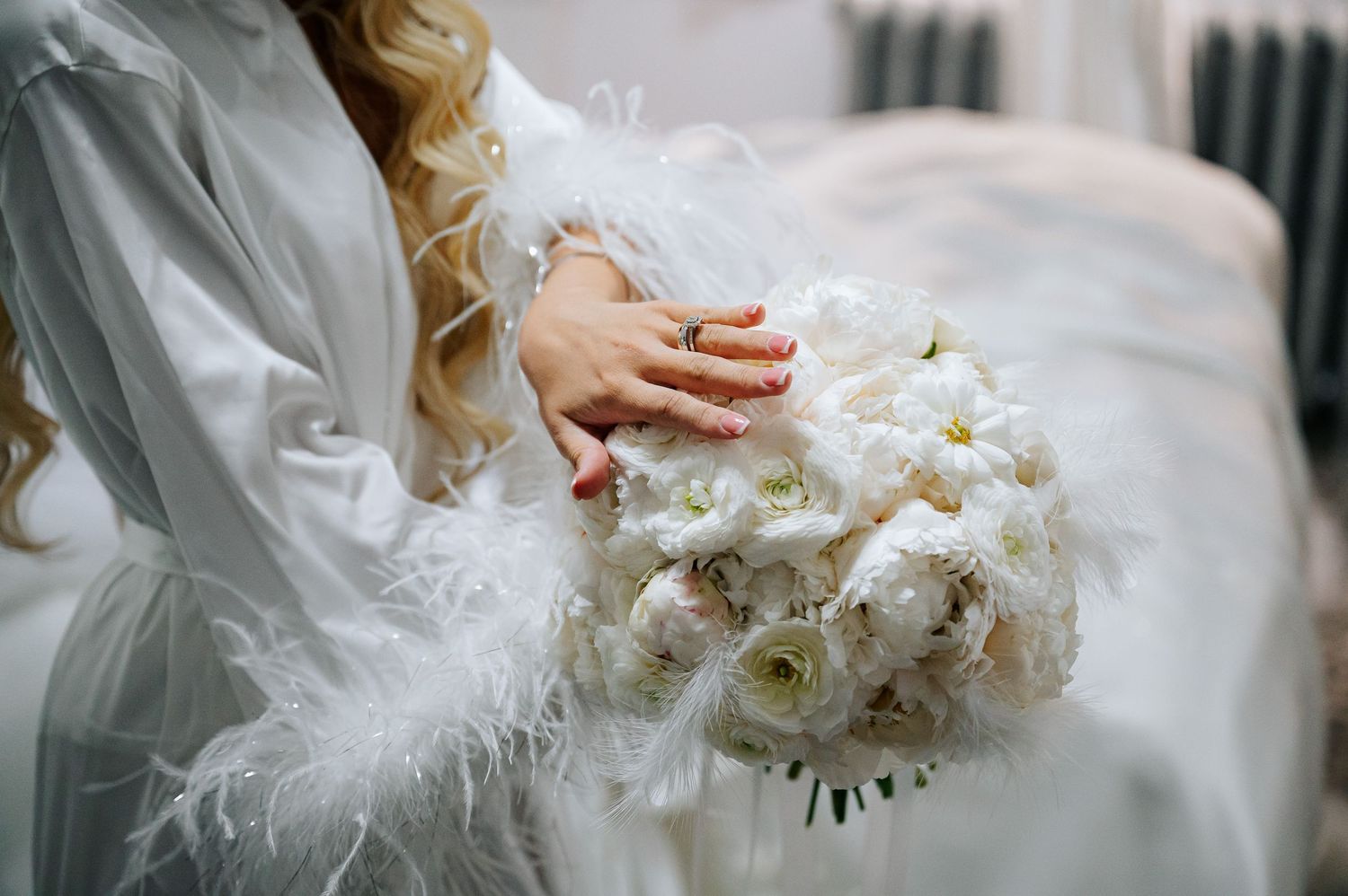 Bride in white feathered robe holds lush white peony and rose bouquet during wedding morning preparations.
