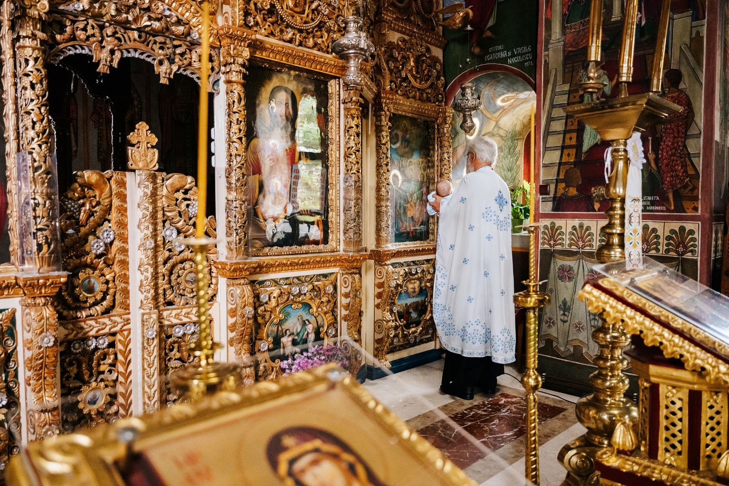 Orthodox priest in white vestments performs a religious ceremony before an ornate golden iconostasis.