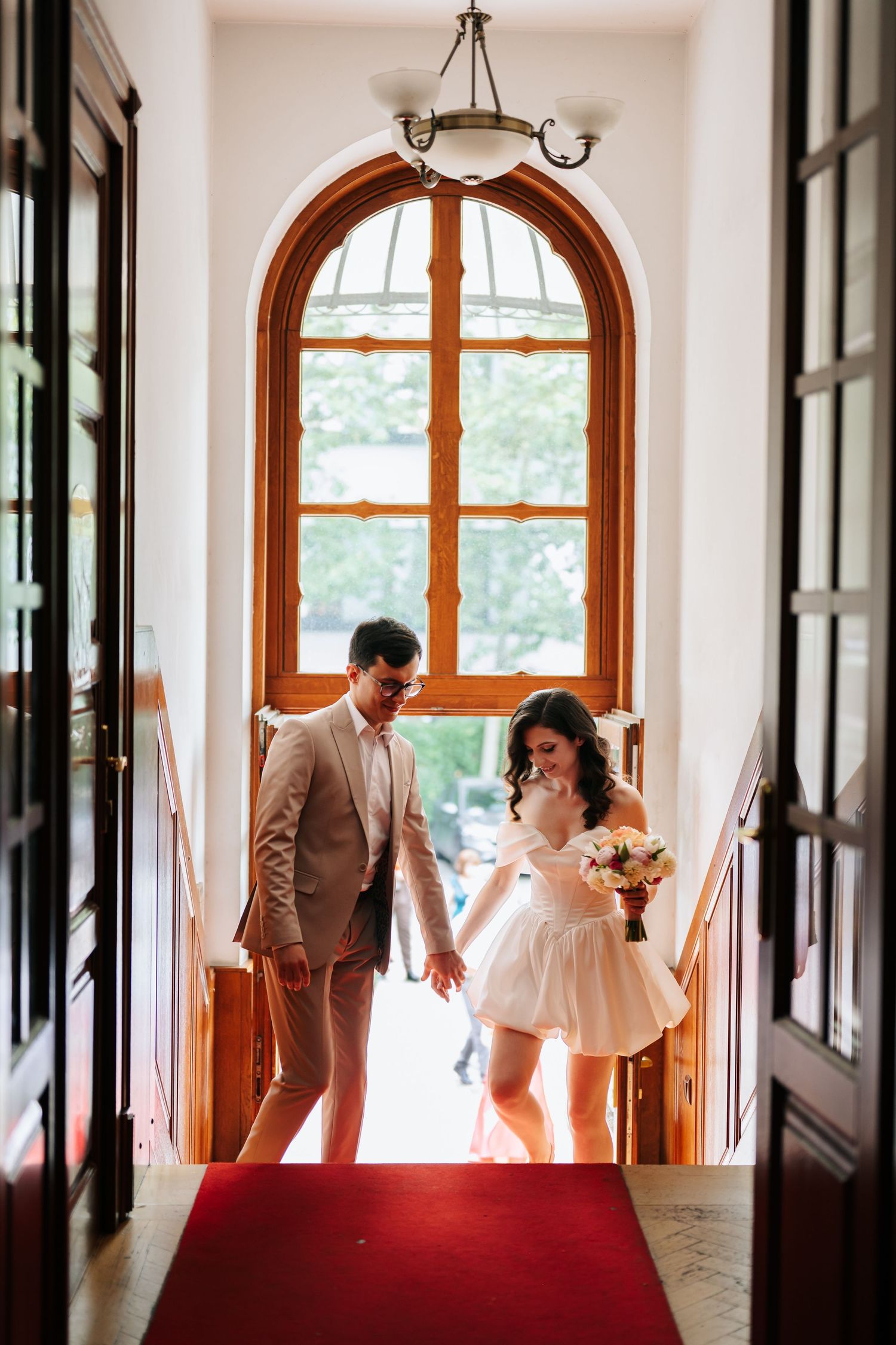 Bride in short white dress and groom in beige suit enter through arched wooden doors on red carpet.