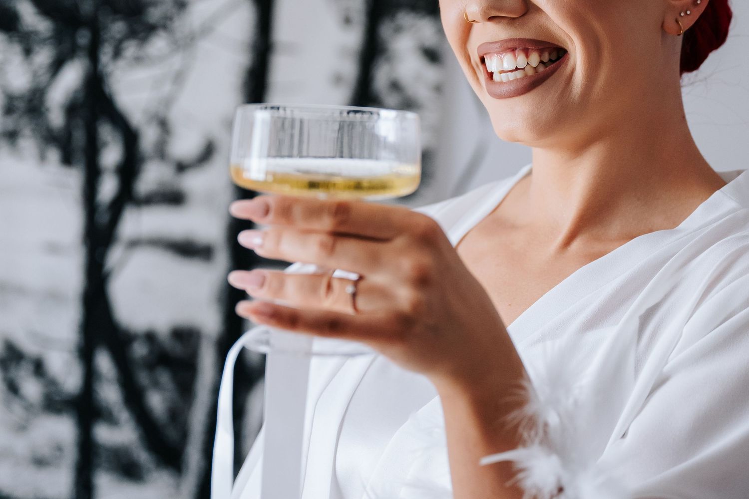 Smiling woman in white robe holding a glass of white wine, wearing a ring, with a blurred background.
