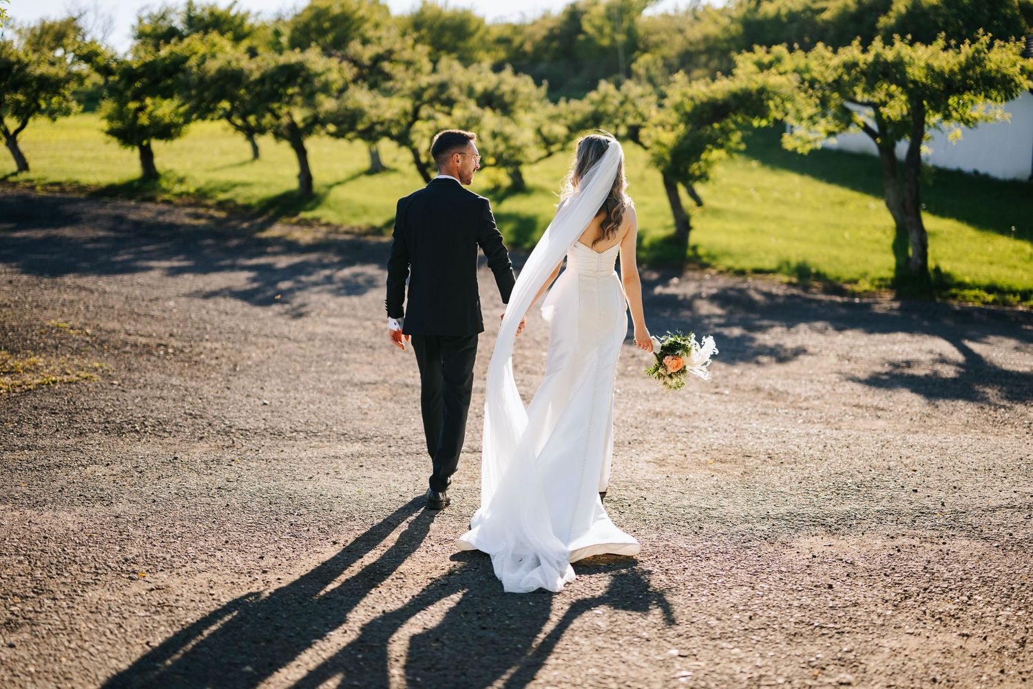 Bride and groom walking hand in hand on a gravel path surrounded by lush green trees on a sunny wedding day.