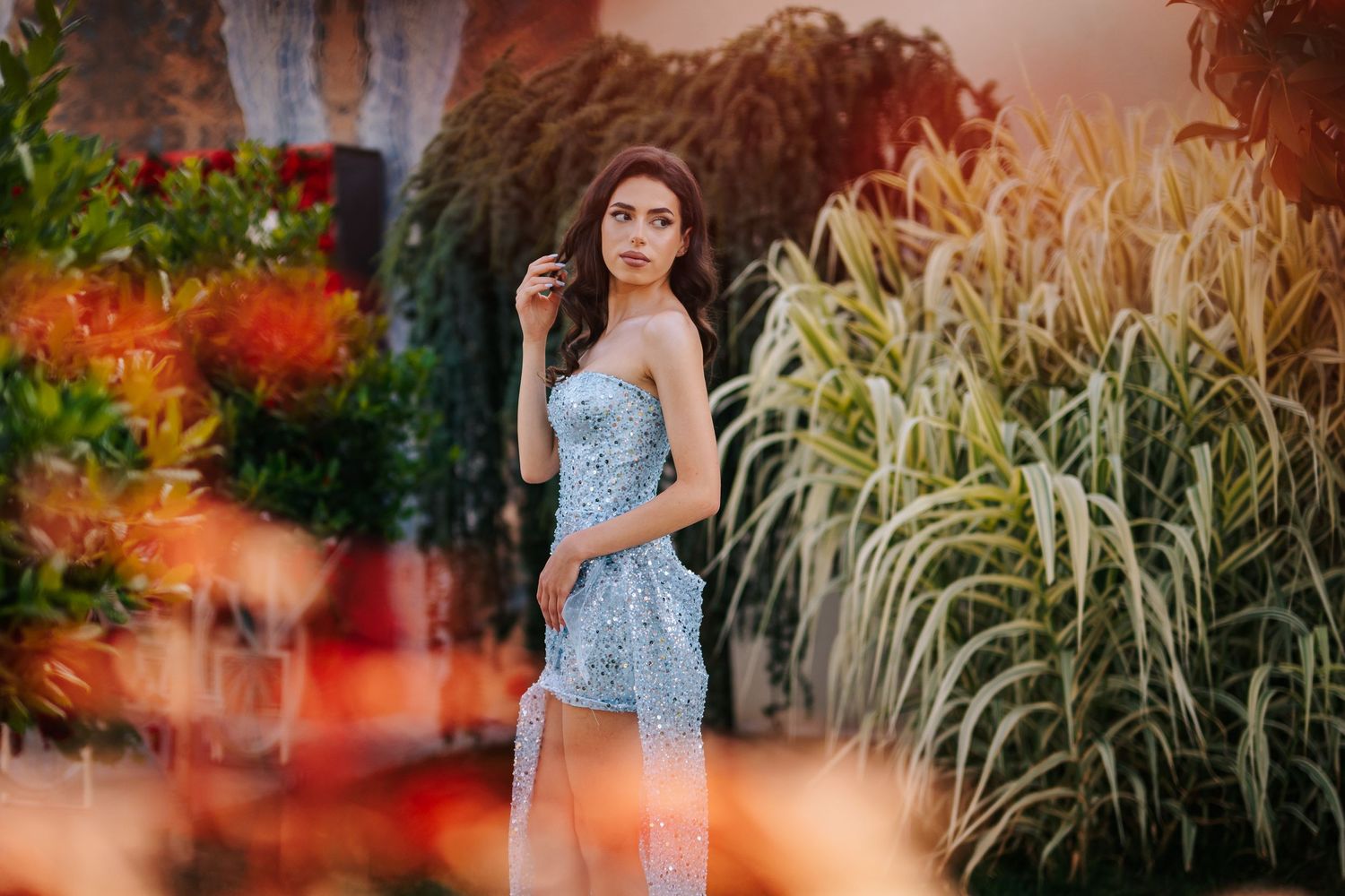 A woman in a blue sequin gown poses gracefully among lush tropical plants and colorful flowers.