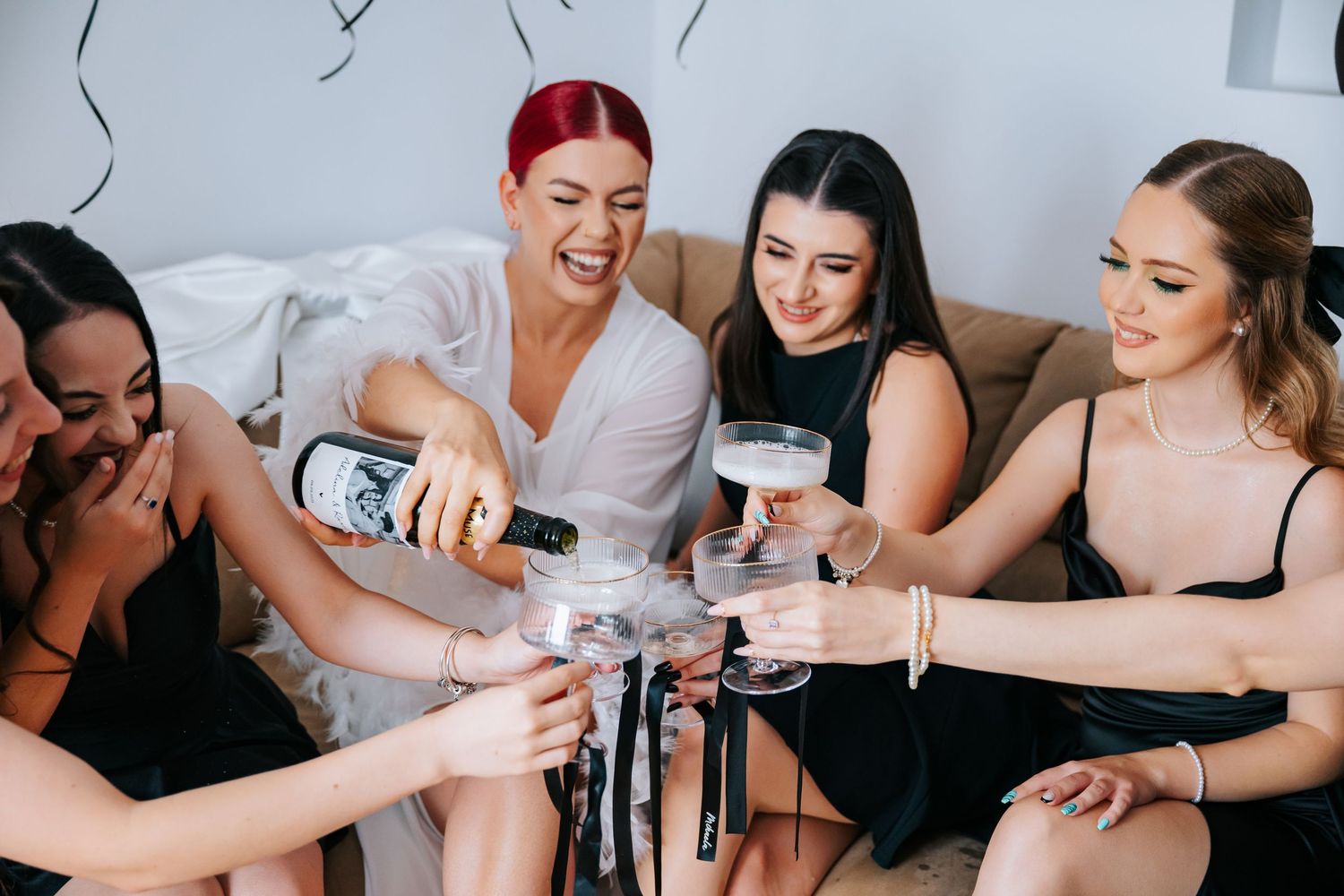 Bride in white robe pouring champagne with bridesmaids in black dresses during a fun bachelorette party celebration.