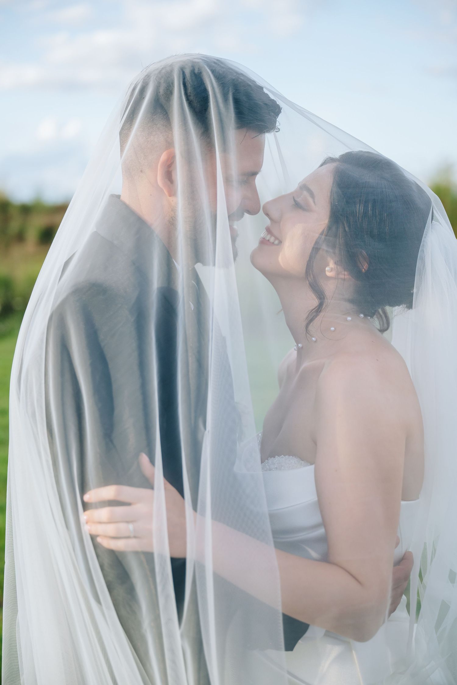 Bride and groom share a romantic moment under a flowing white veil outdoors on their wedding day.